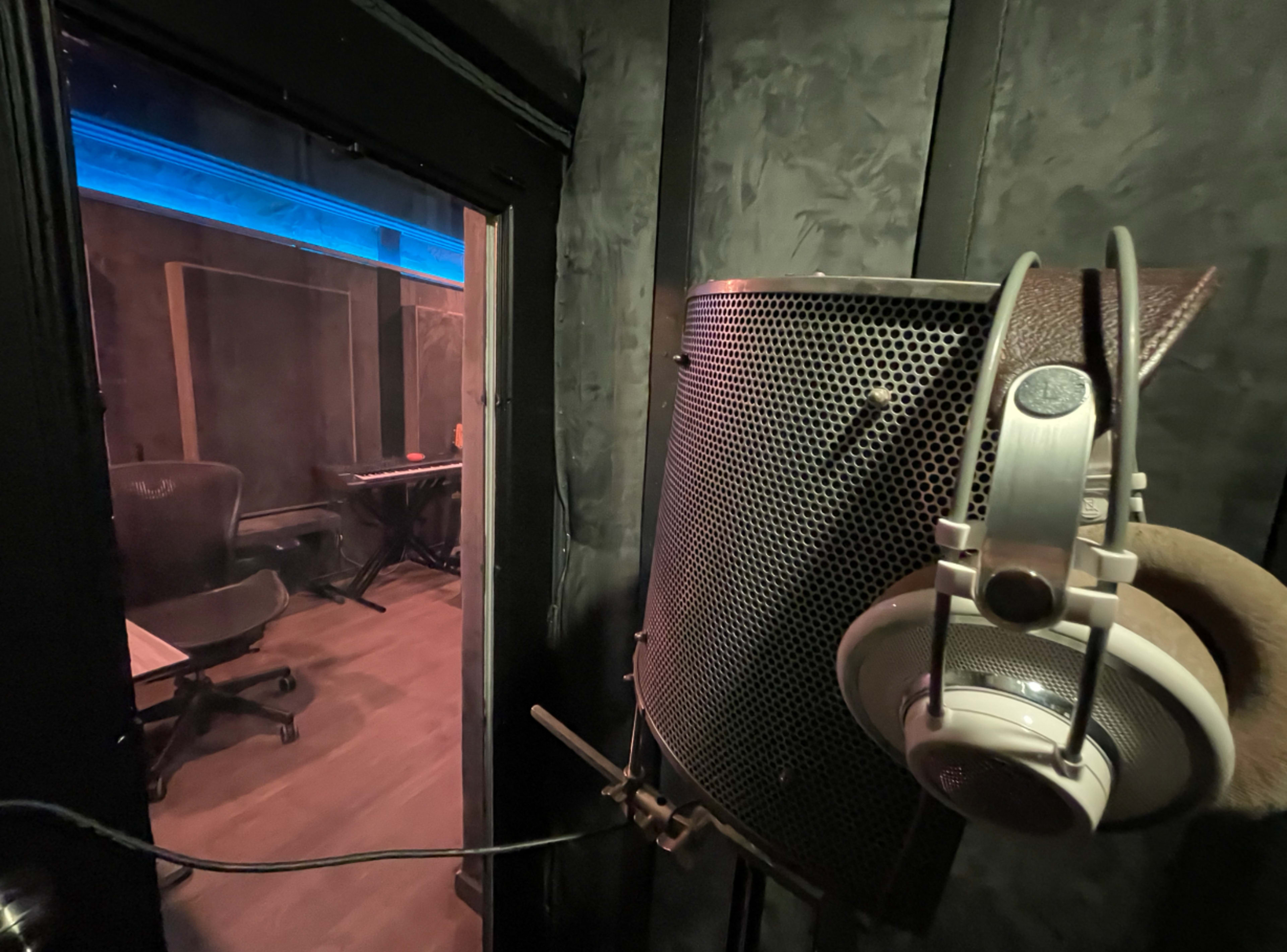 The image shows a recording studio booth with a microphone, headphones, and a view of a room featuring a keyboard and a chair.
