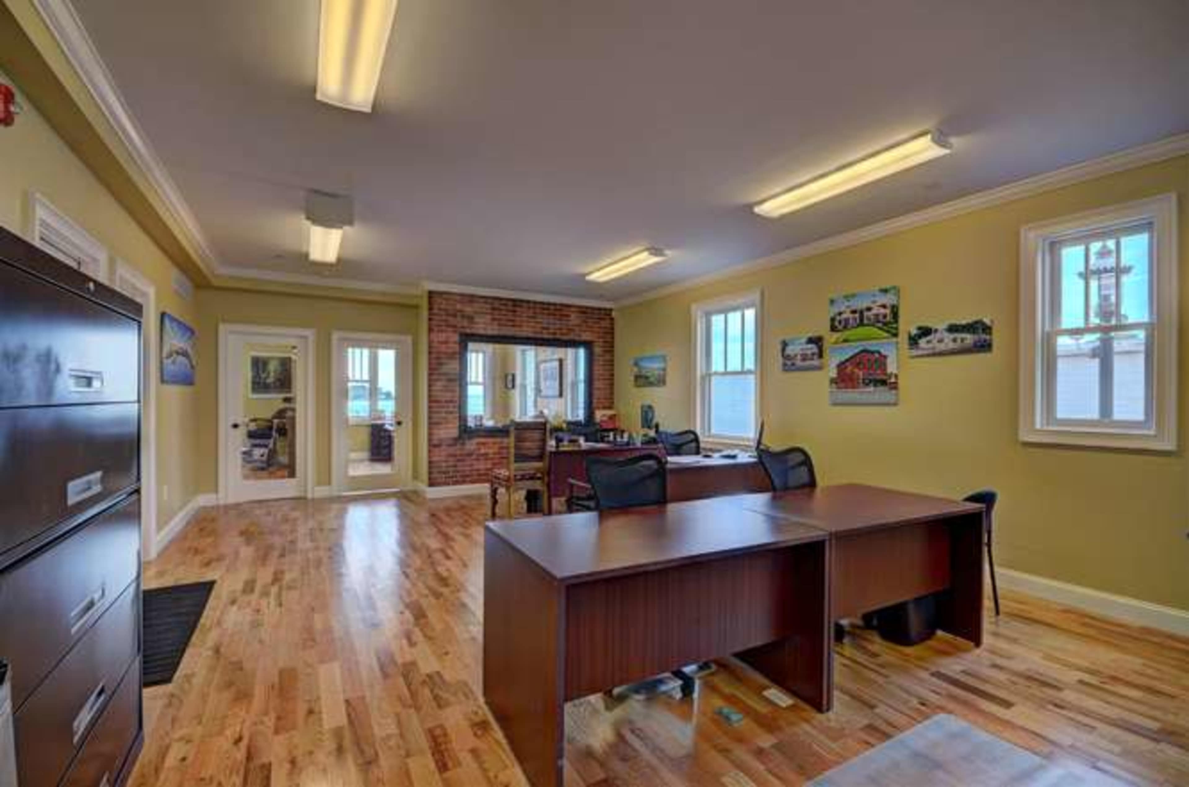 The image depicts a modern office space with wooden floors, brick accent walls, and multiple windows allowing natural light, furnished with desks and filing cabinets.