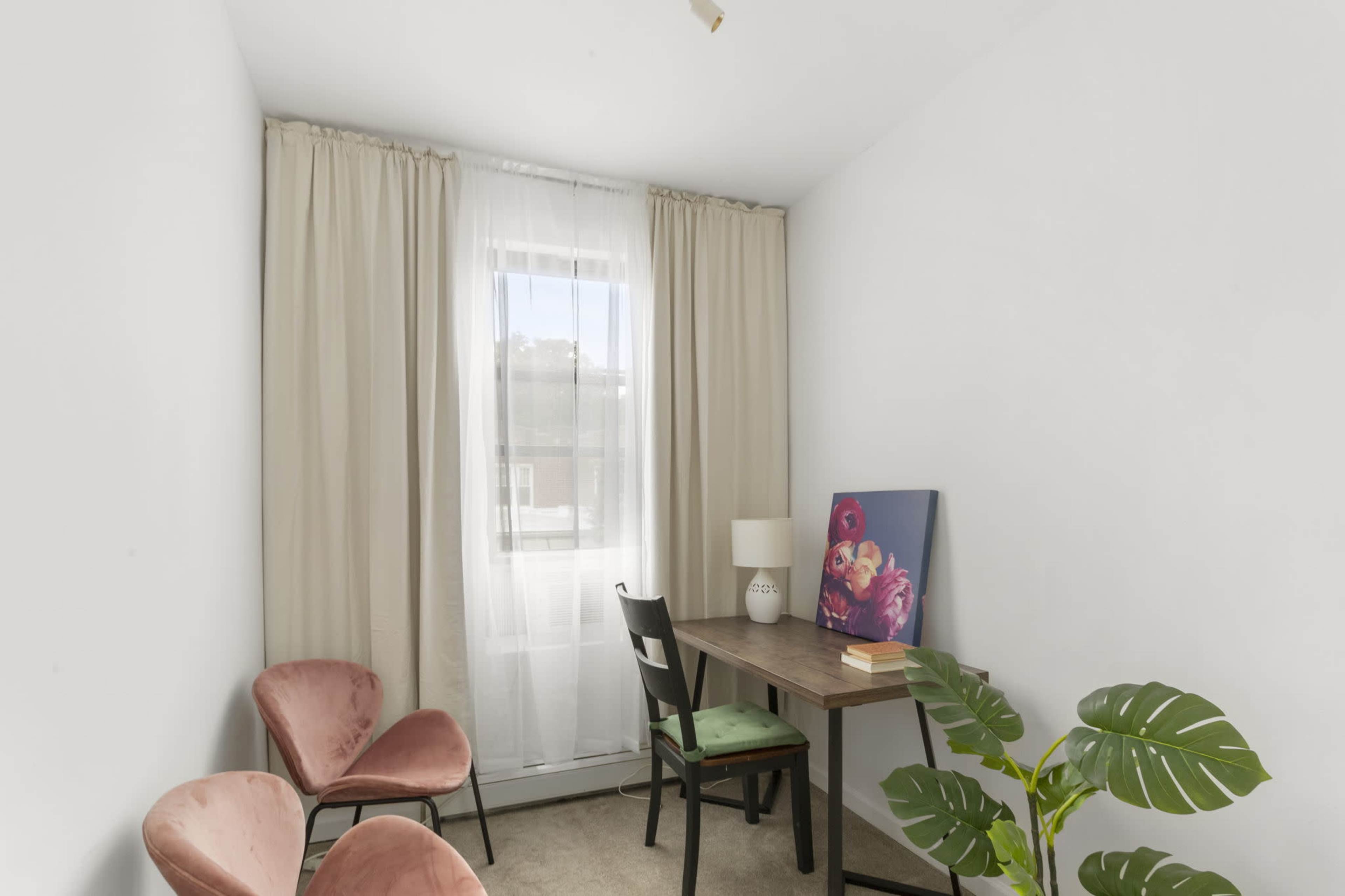 A small, minimalist room features a desk with a lamp, two pink chairs, and large curtains covering a window.