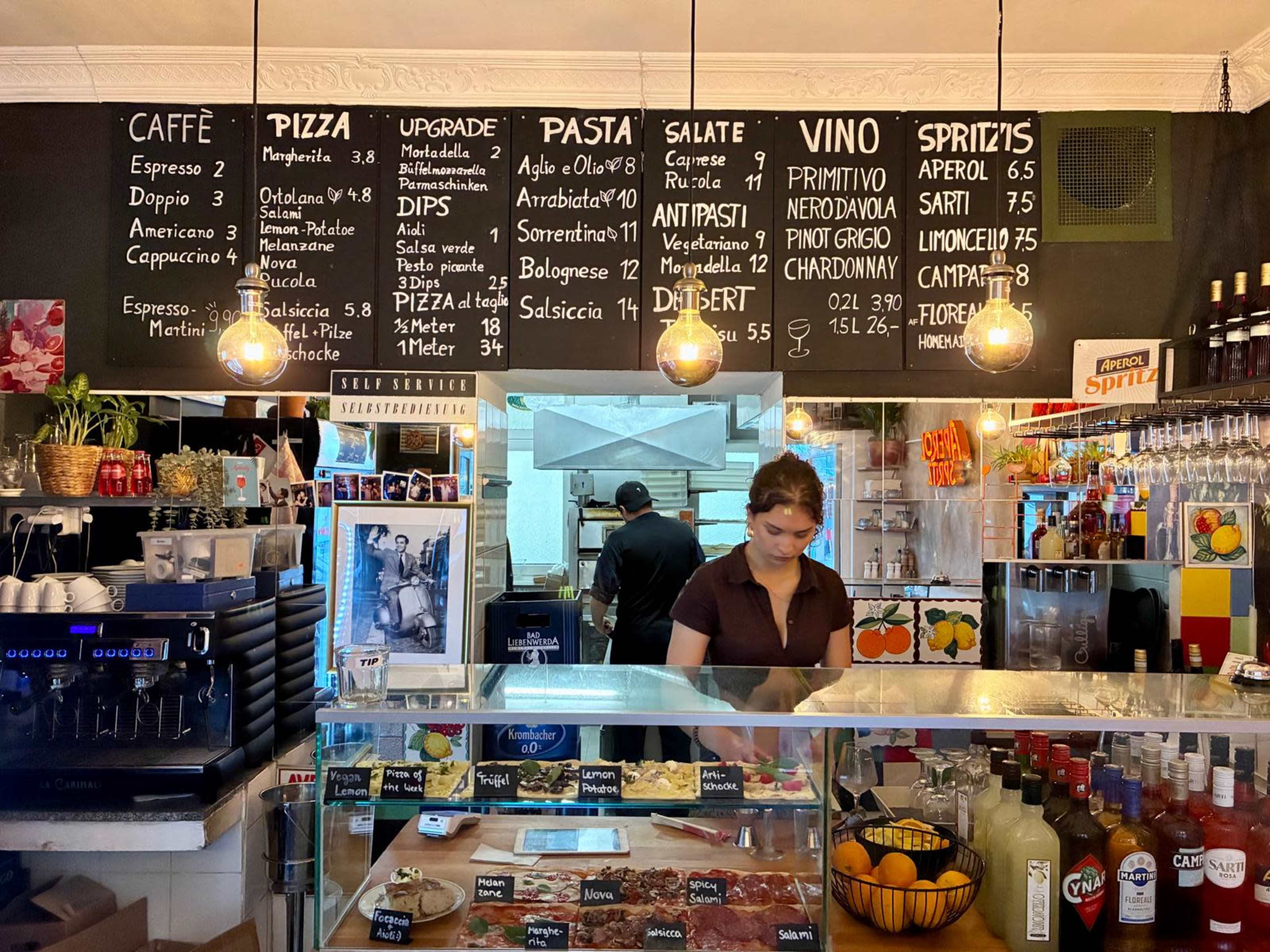 The image depicts a bustling café interior with a menu board featuring various Italian dishes and drinks, while a staff member prepares orders behind the counter.