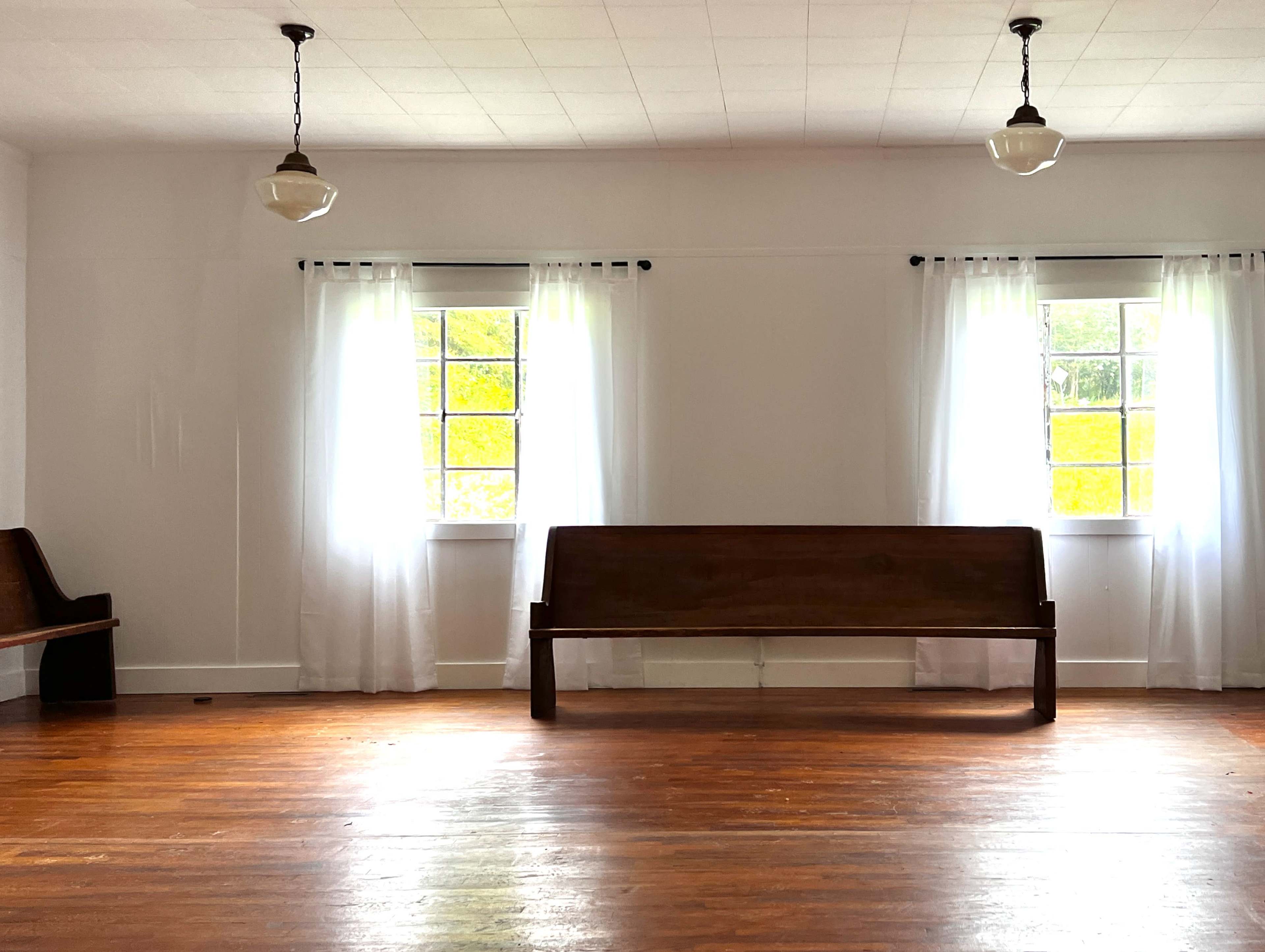 The interior of a sparsely furnished room features two wooden benches along the walls and large windows with sheer white curtains letting in natural light.