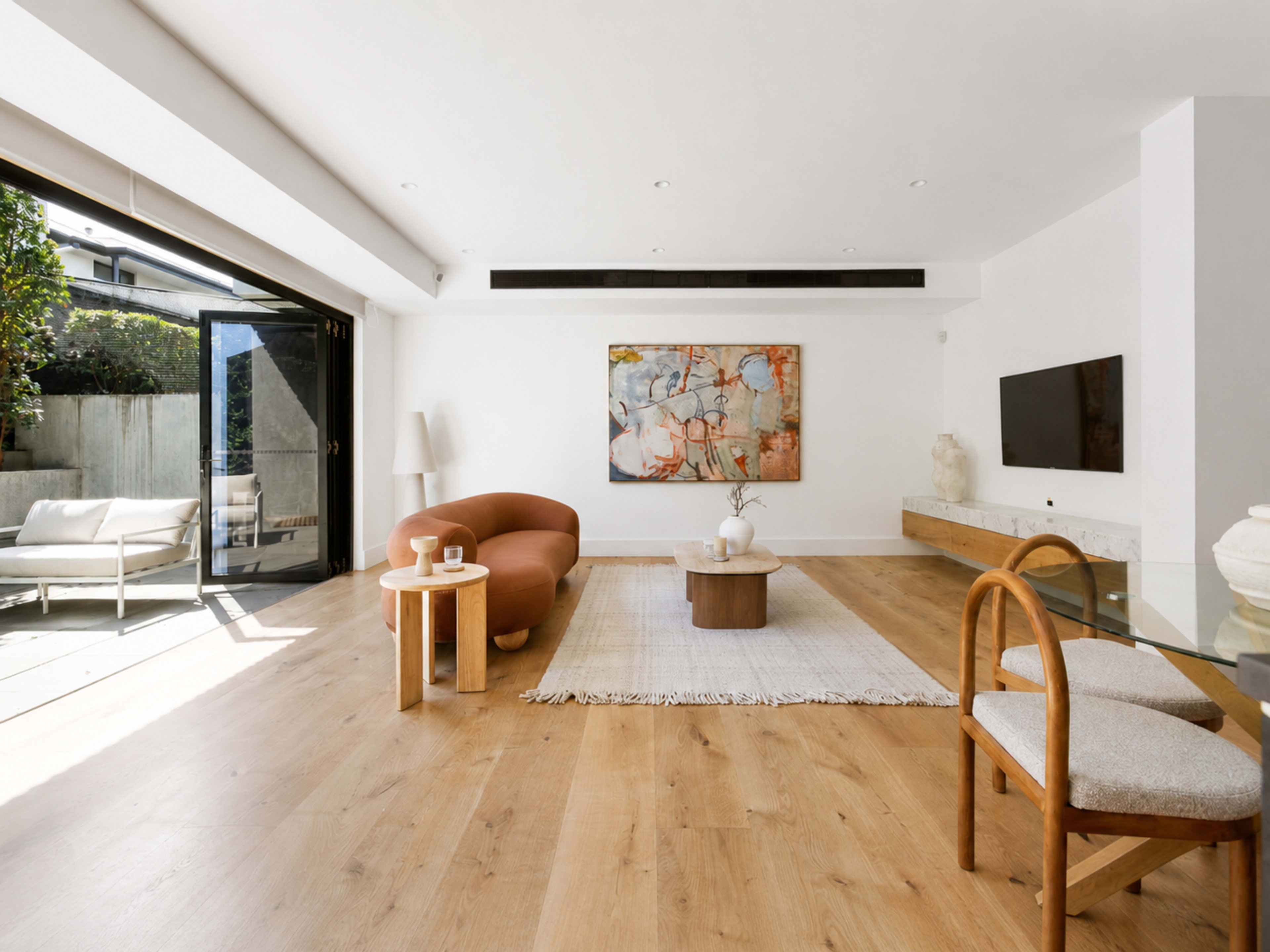 The image shows a modern living room with a curved orange sofa, a light wooden floor, and large glass doors leading to an outdoor space.
