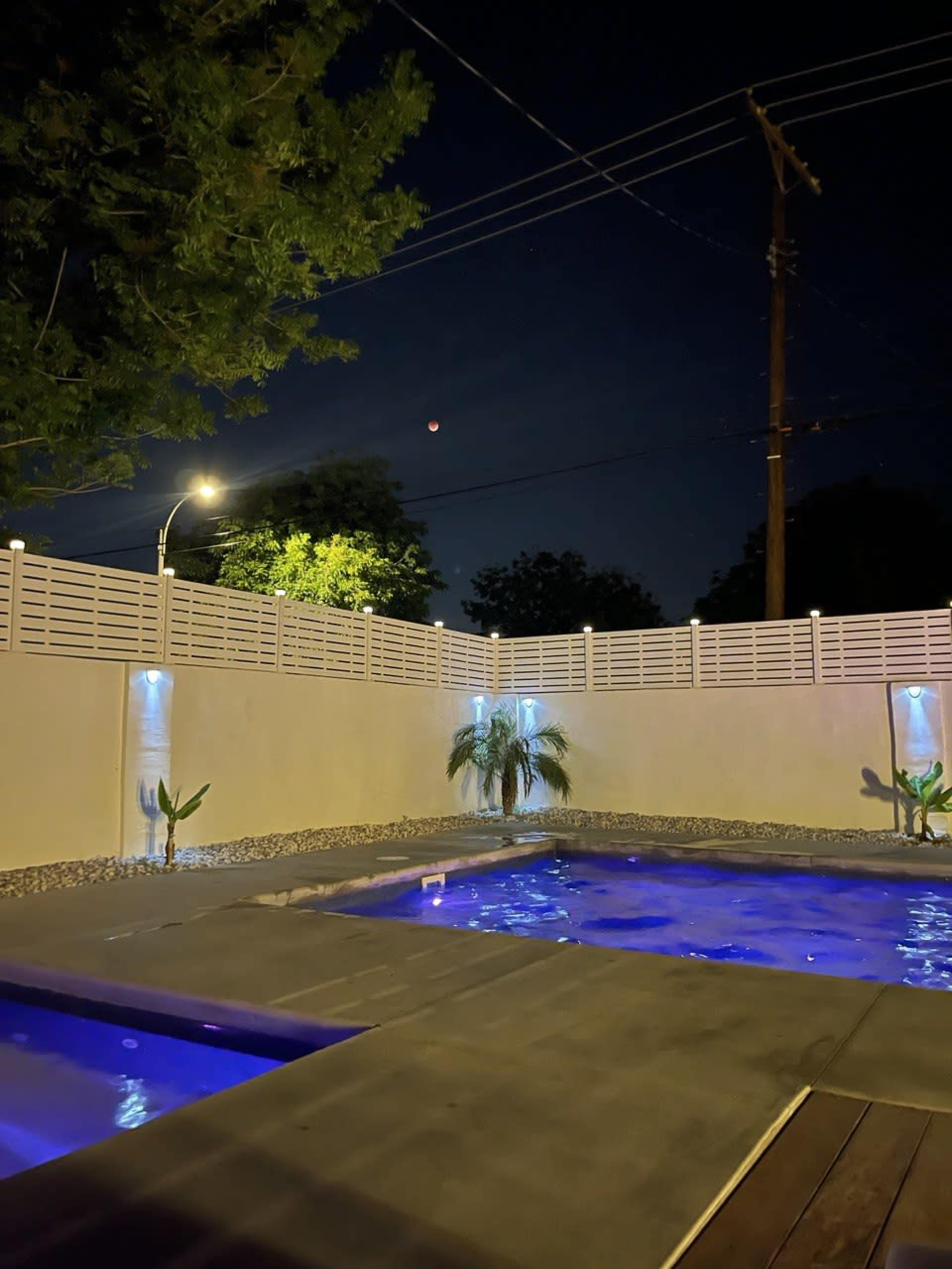 A backyard with two illuminated swimming pools under a night sky, featuring a partial view of the moon and surrounding greenery.