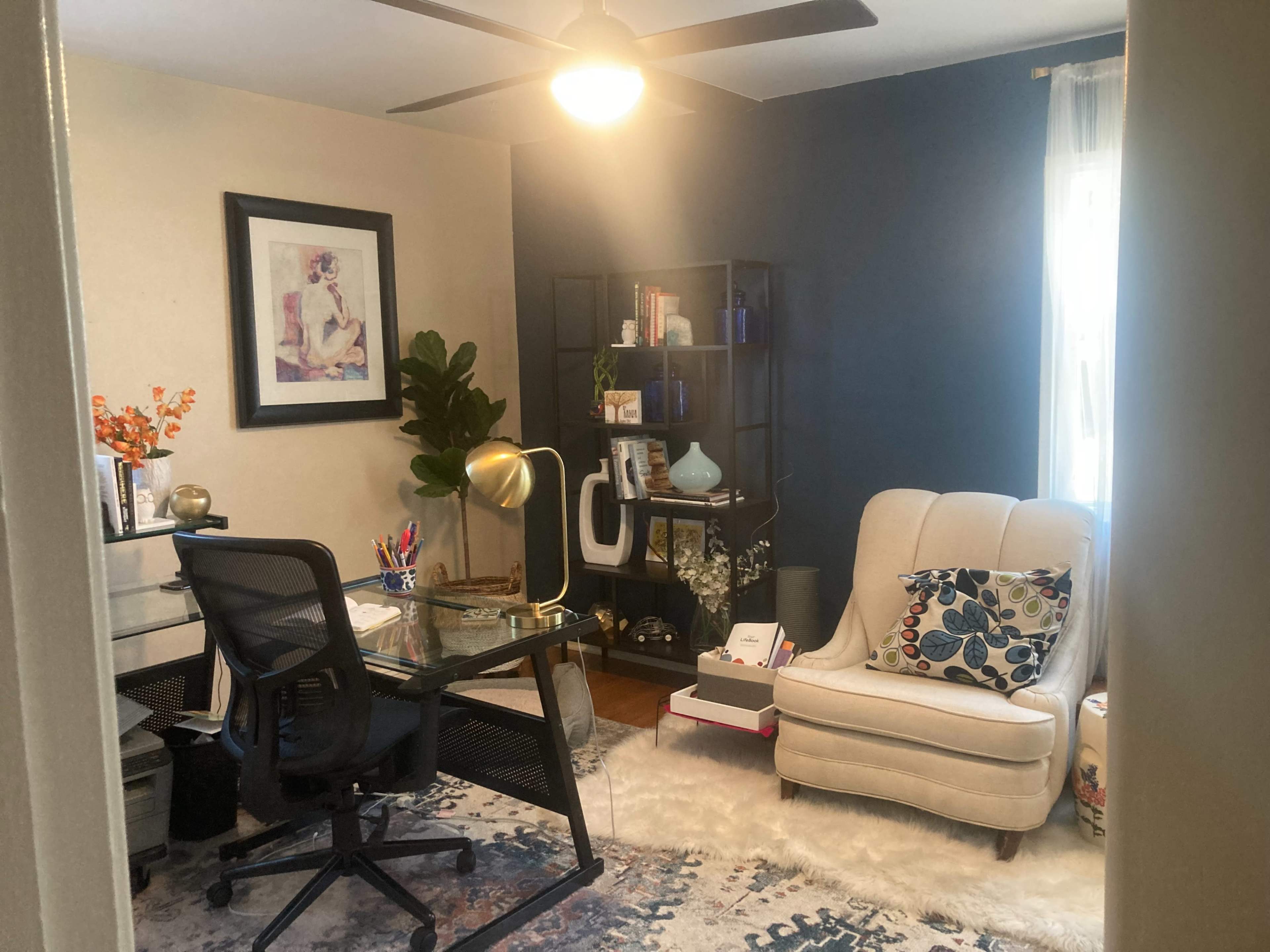 A home office with a desk and chair, a light-colored armchair, shelves filled with various items, and a large rug on the floor.