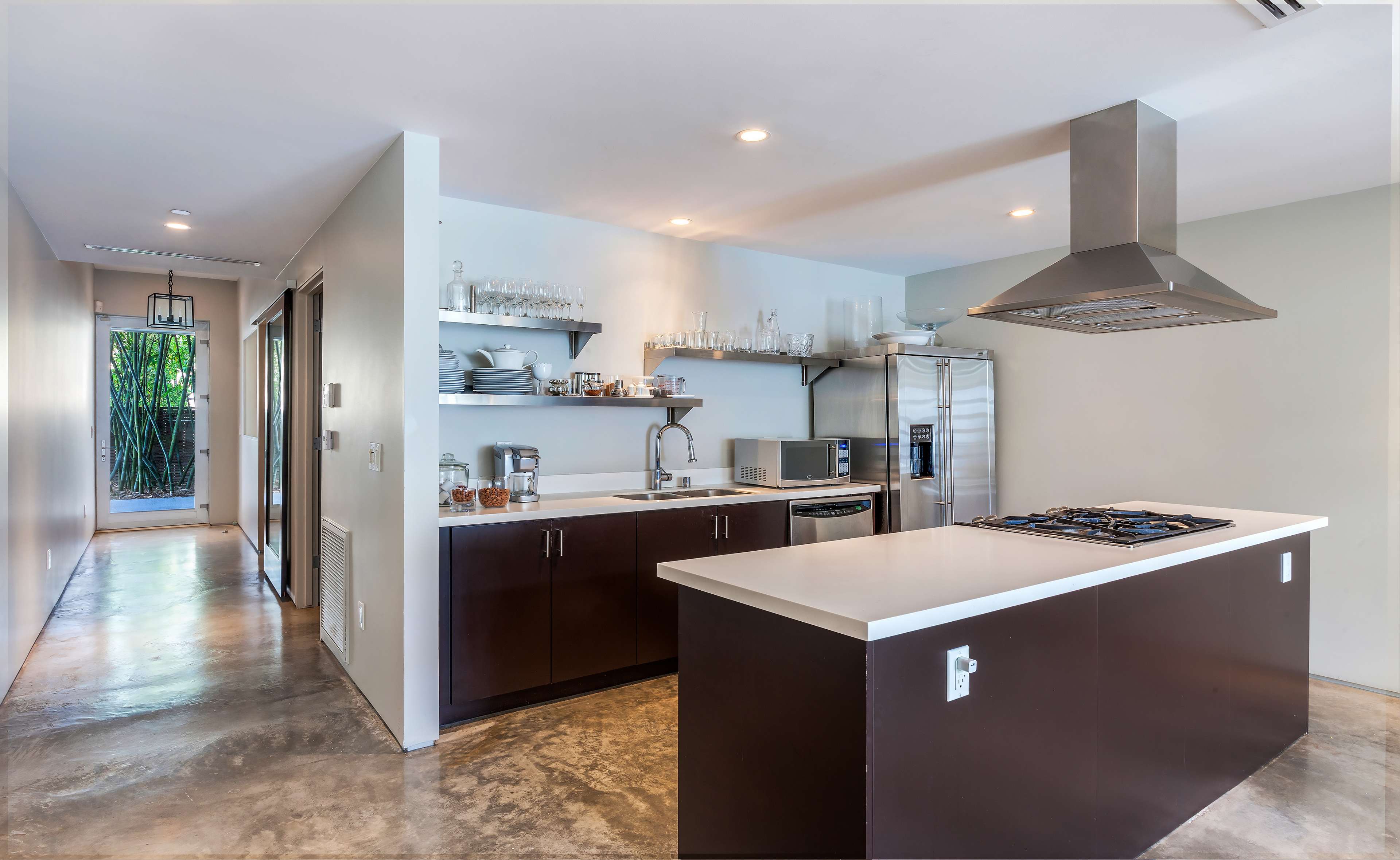 The image shows a modern kitchen with an island, stainless steel appliances, and open shelving, along with a hallway leading to an entrance.