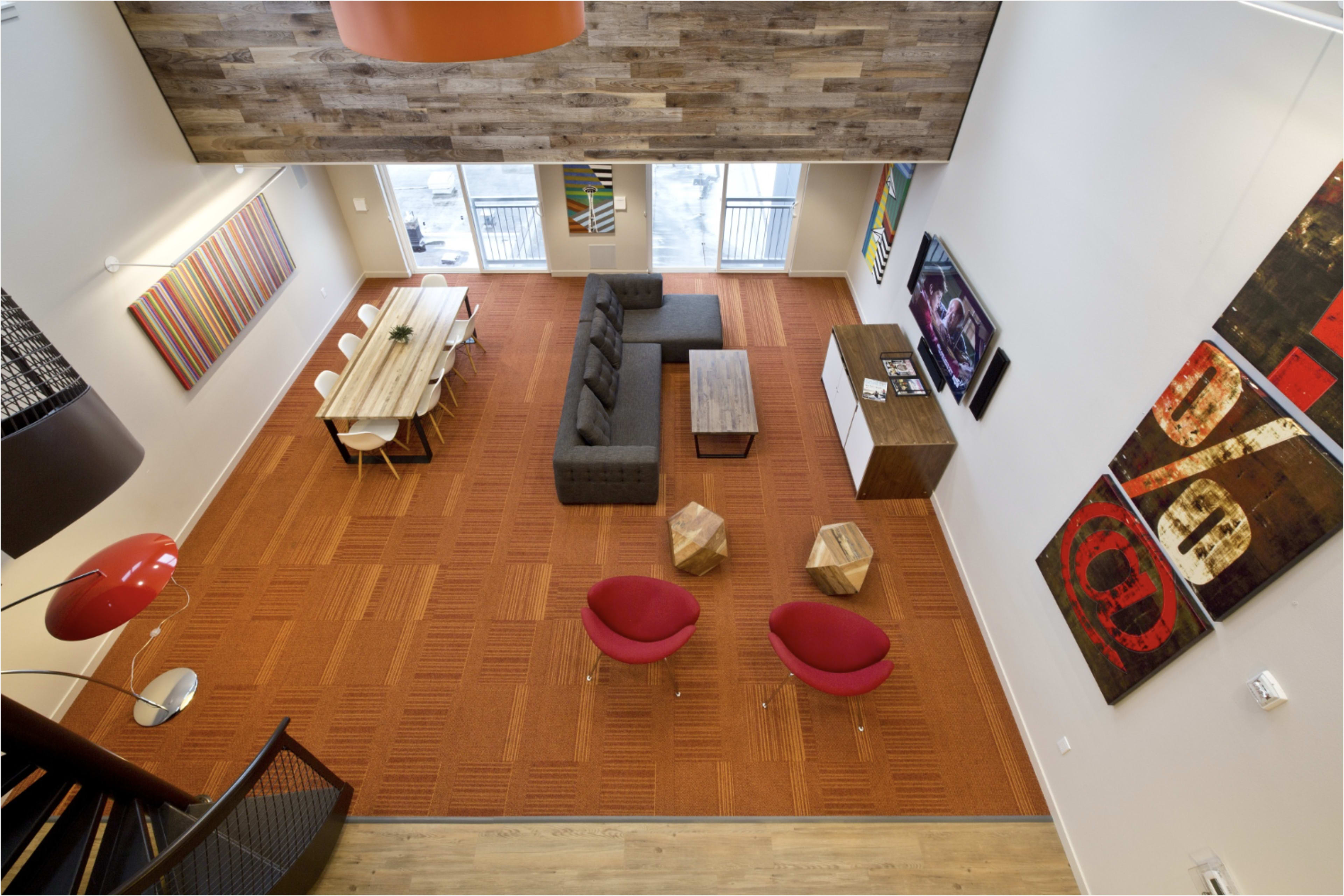 The image shows a spacious modern living area featuring a rectangular dining table, a sectional sofa, two red chairs, and various wall artworks, all set on an orange carpet.