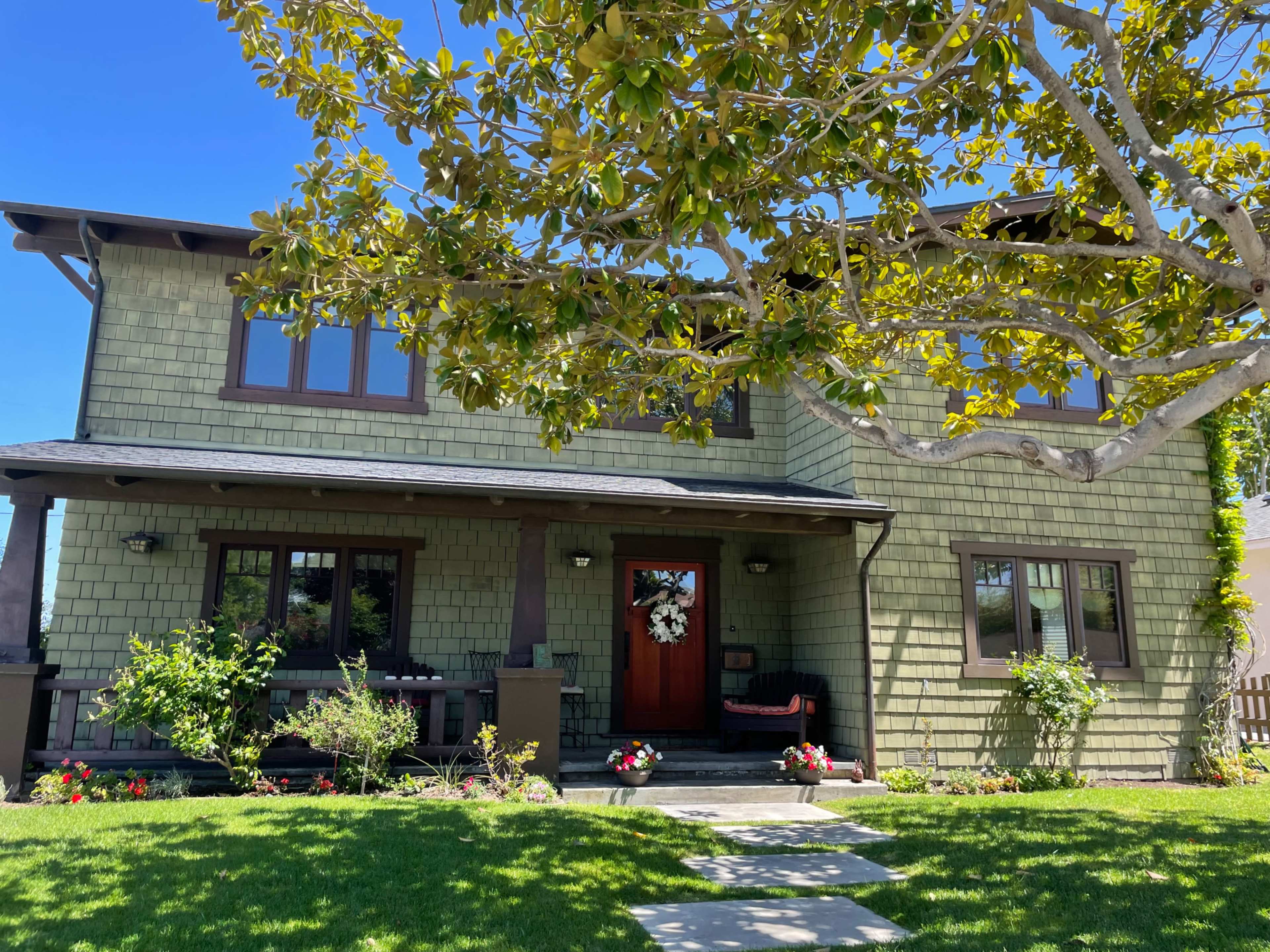 A two-story, green-shingle house with a front porch and a wreath on the door is surrounded by a well-maintained lawn and flowering plants.