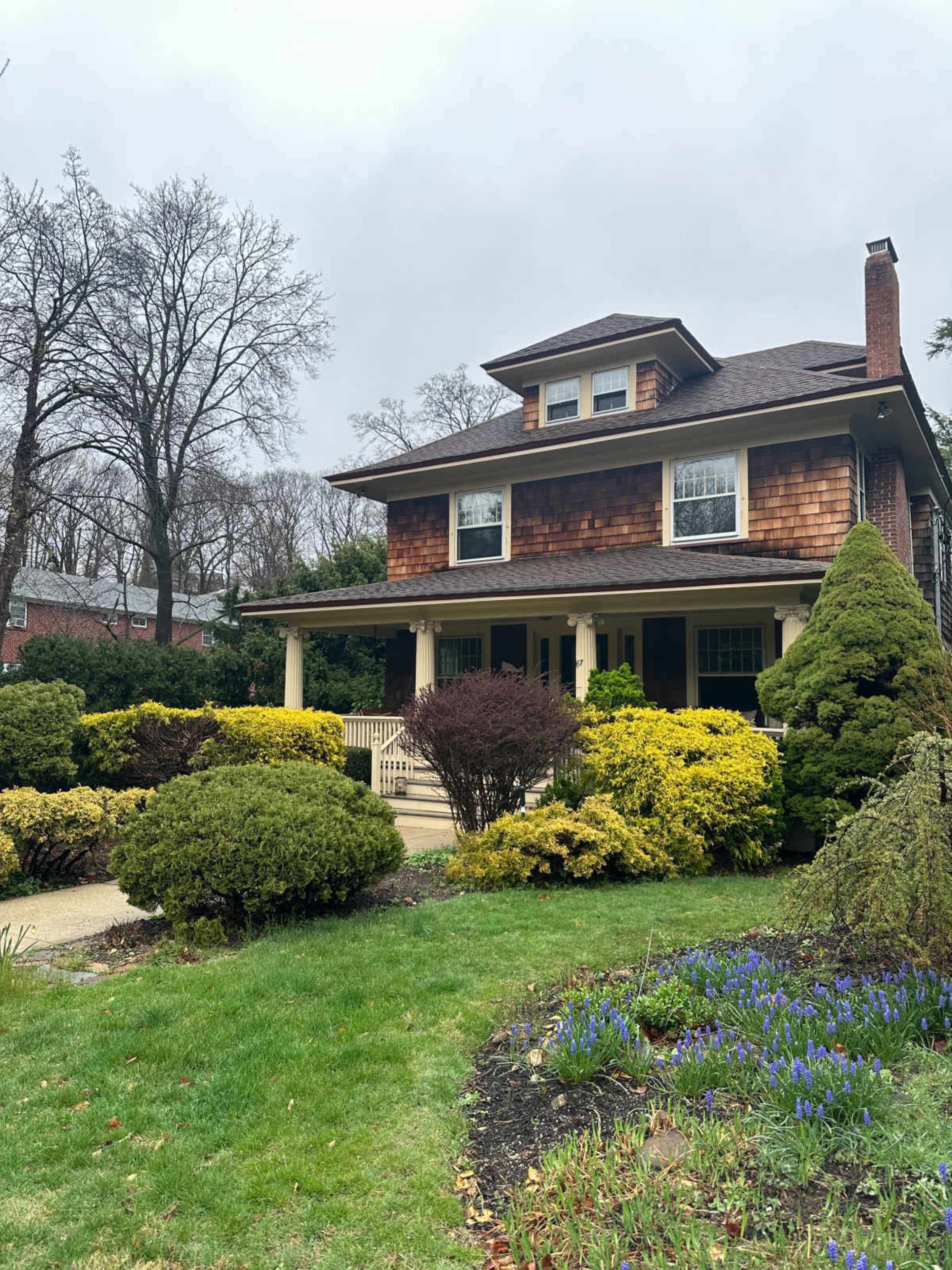 The image shows a two-story house with a shingle-style facade surrounded by well-maintained shrubs and flowers in a green lawn.
