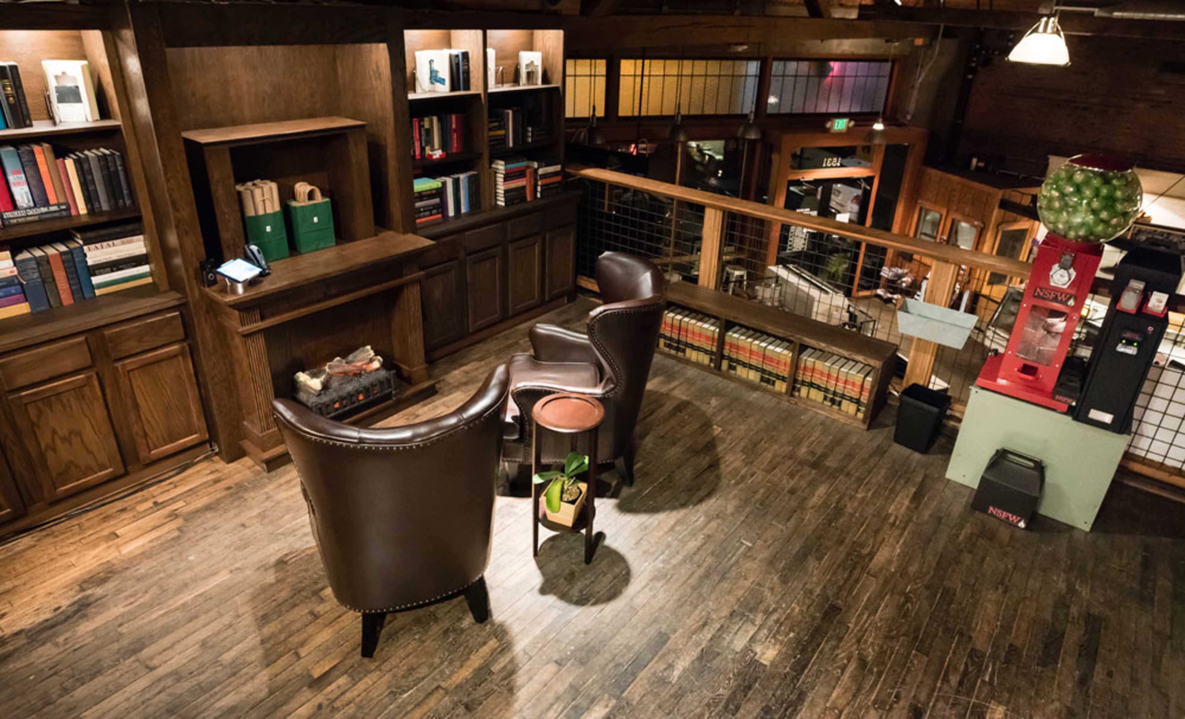 The image shows a cozy reading nook with two leather chairs facing each other, surrounded by wooden bookshelves, and overlooking a spacious interior with wooden flooring and additional seating areas.