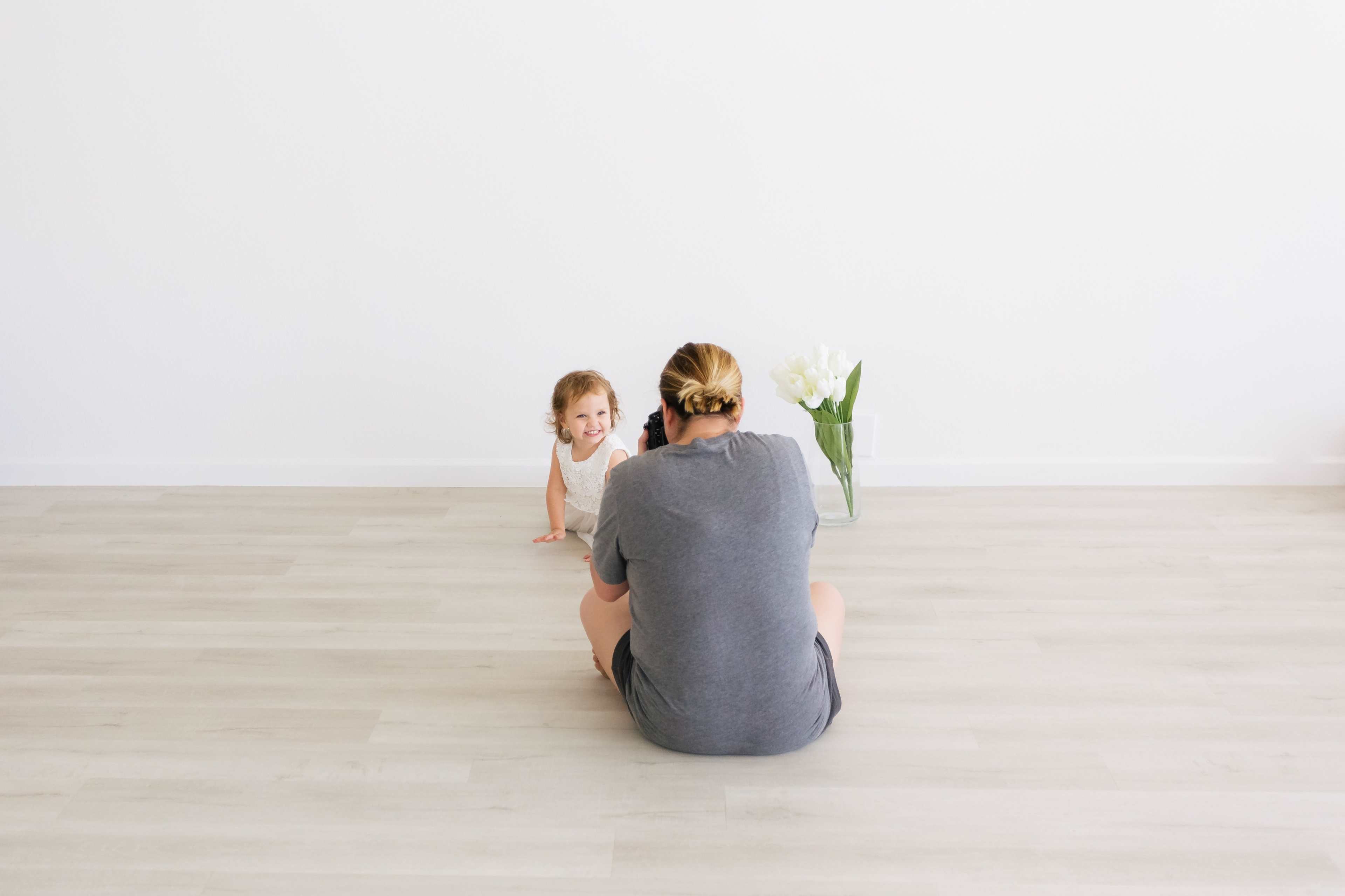 A child crawls toward a person sitting on the floor in a minimalist room with a vase of flowers.