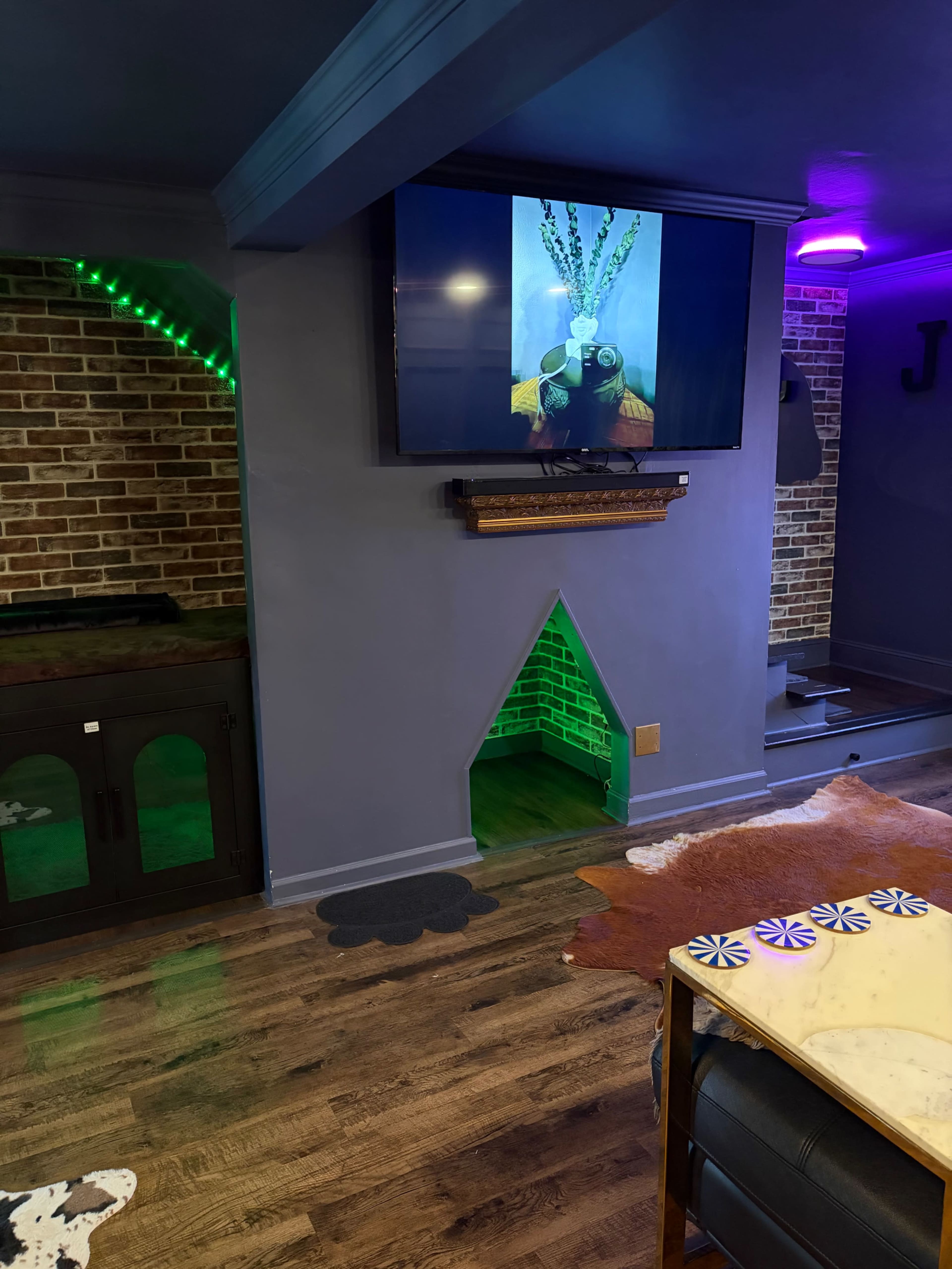 The image shows a cozy interior space featuring a television mounted on a wall, a brick accent wall, and a small archway leading to a hidden area, with decorative lighting adding color to the room.