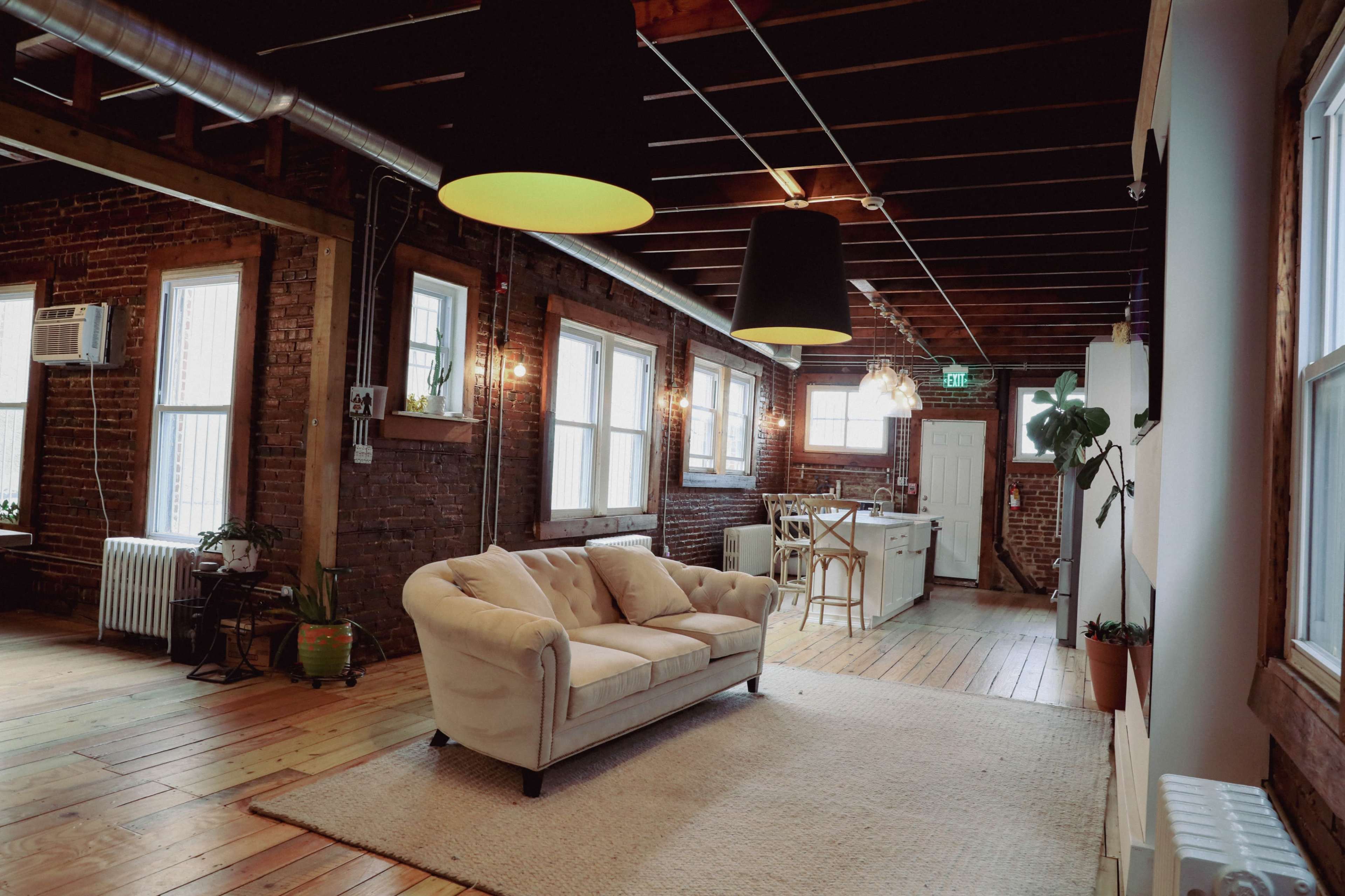 A spacious room with exposed brick walls, featuring a cream-colored couch on a light rug, with large windows and a kitchen area visible in the background.