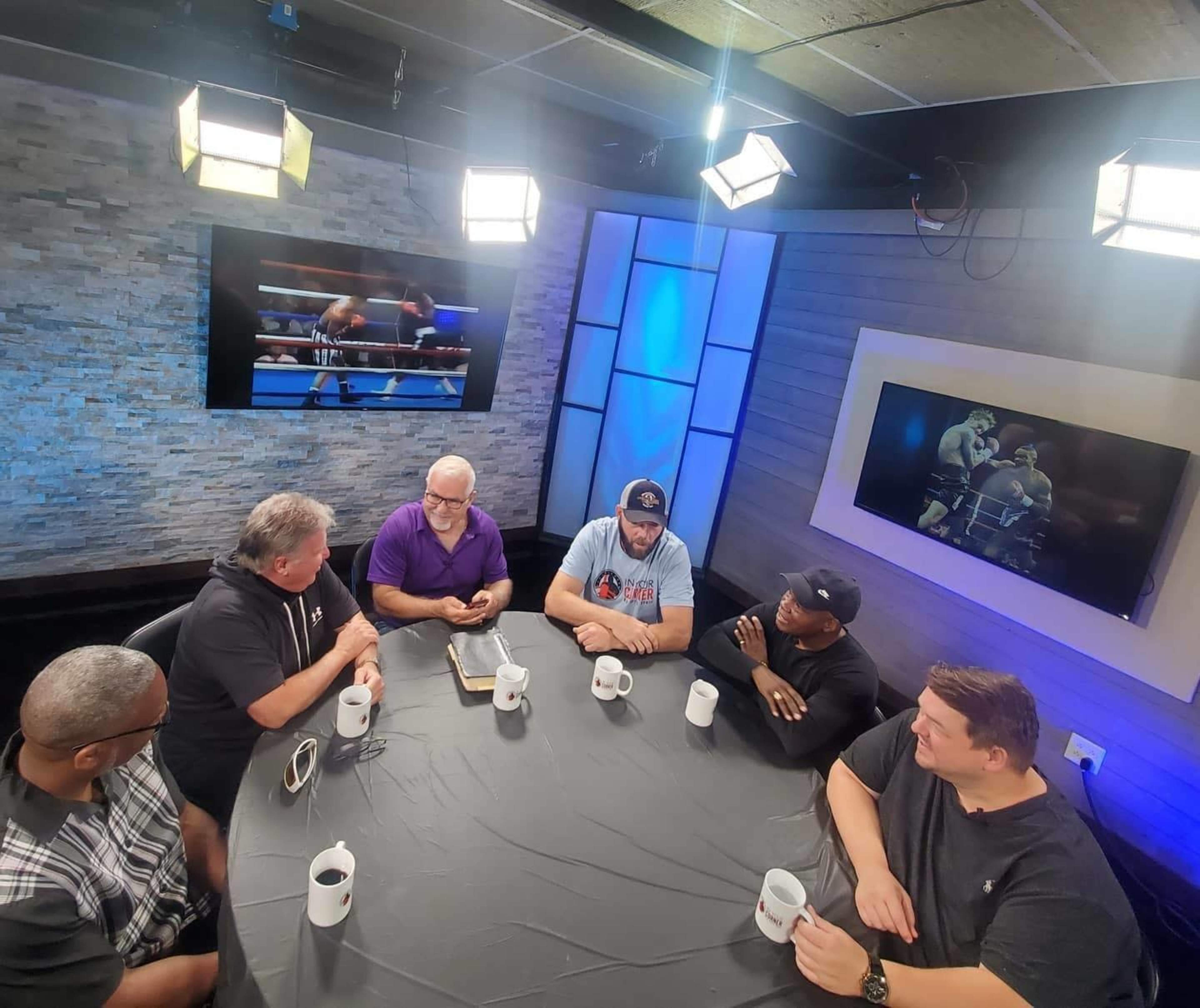 A group of six people sits around a circular table in a studio, discussing a boxing match while screens display fight highlights.