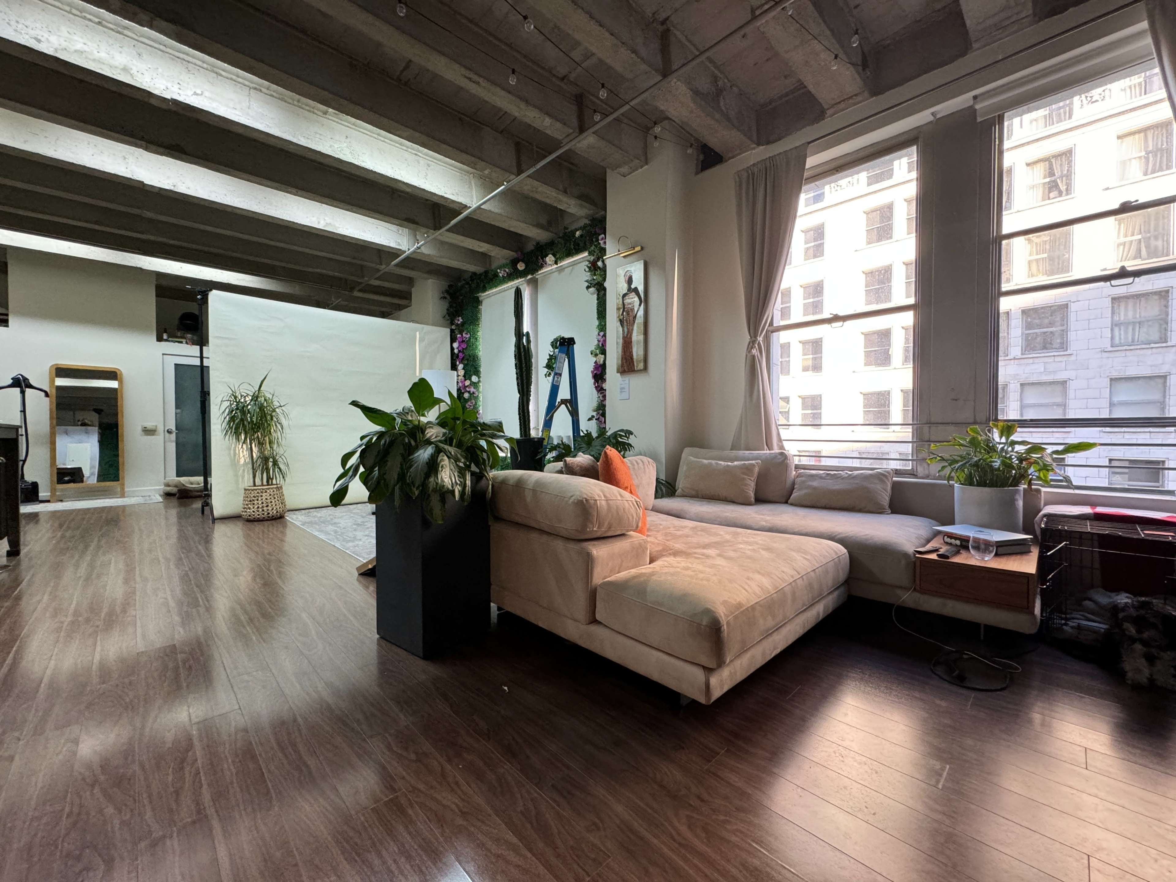 A bright open-plan living space featuring a beige sectional couch, large windows with city views, and greenery throughout the room.