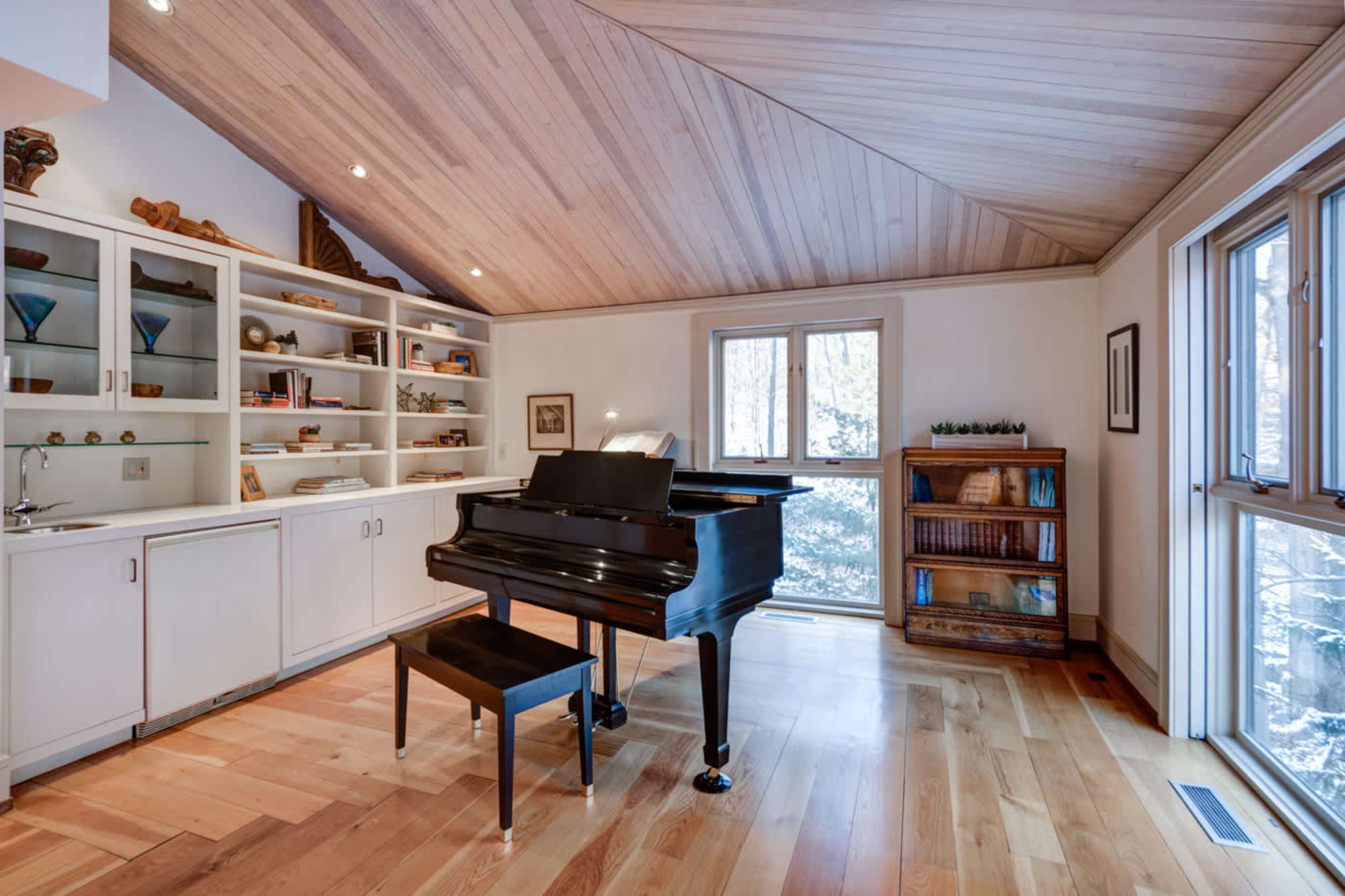 A spacious room featuring a black grand piano, a wooden floor, built-in shelves with various items, and a large window with natural light.