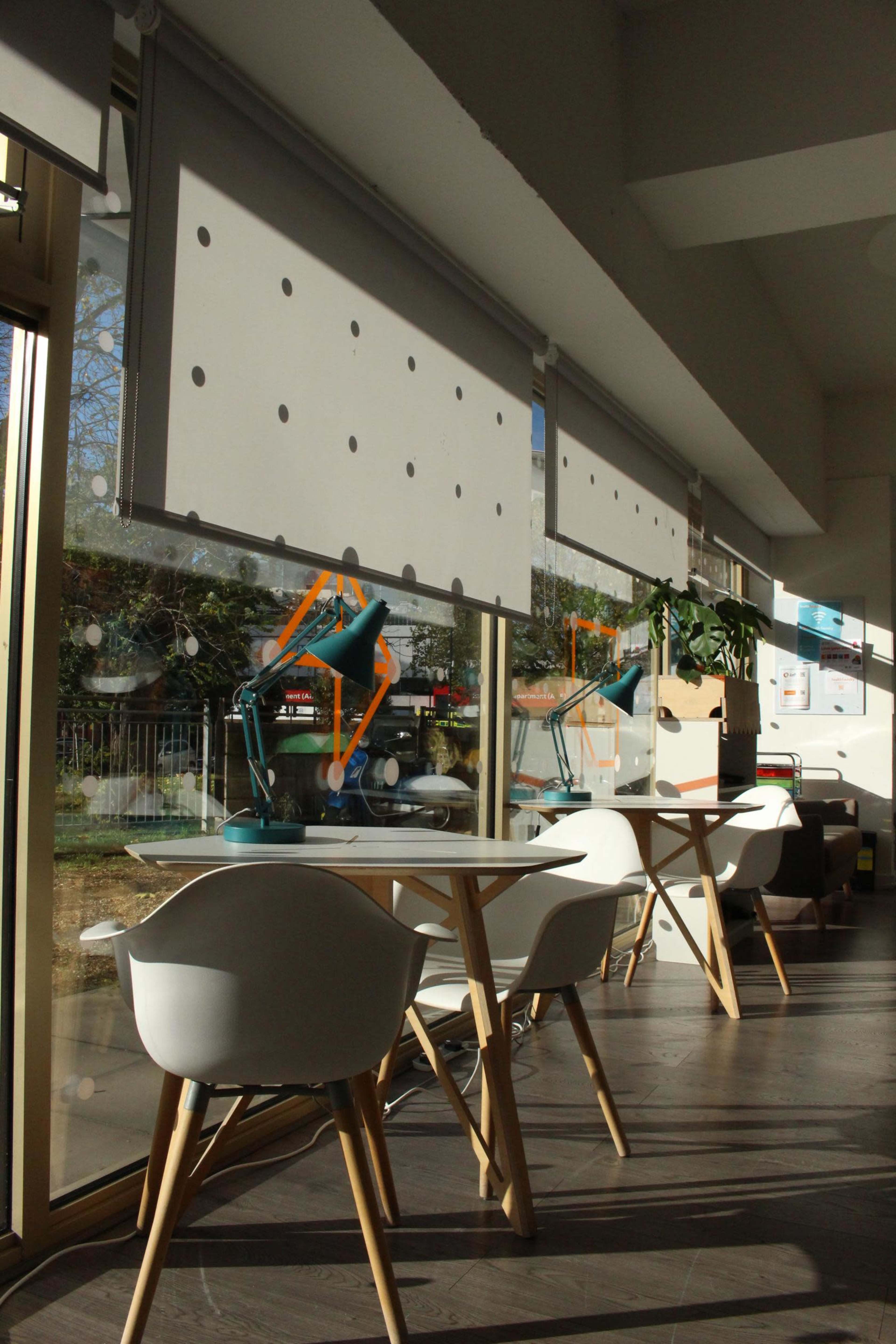 The image shows a bright interior space with white chairs and tables near large windows covered by roller shades.