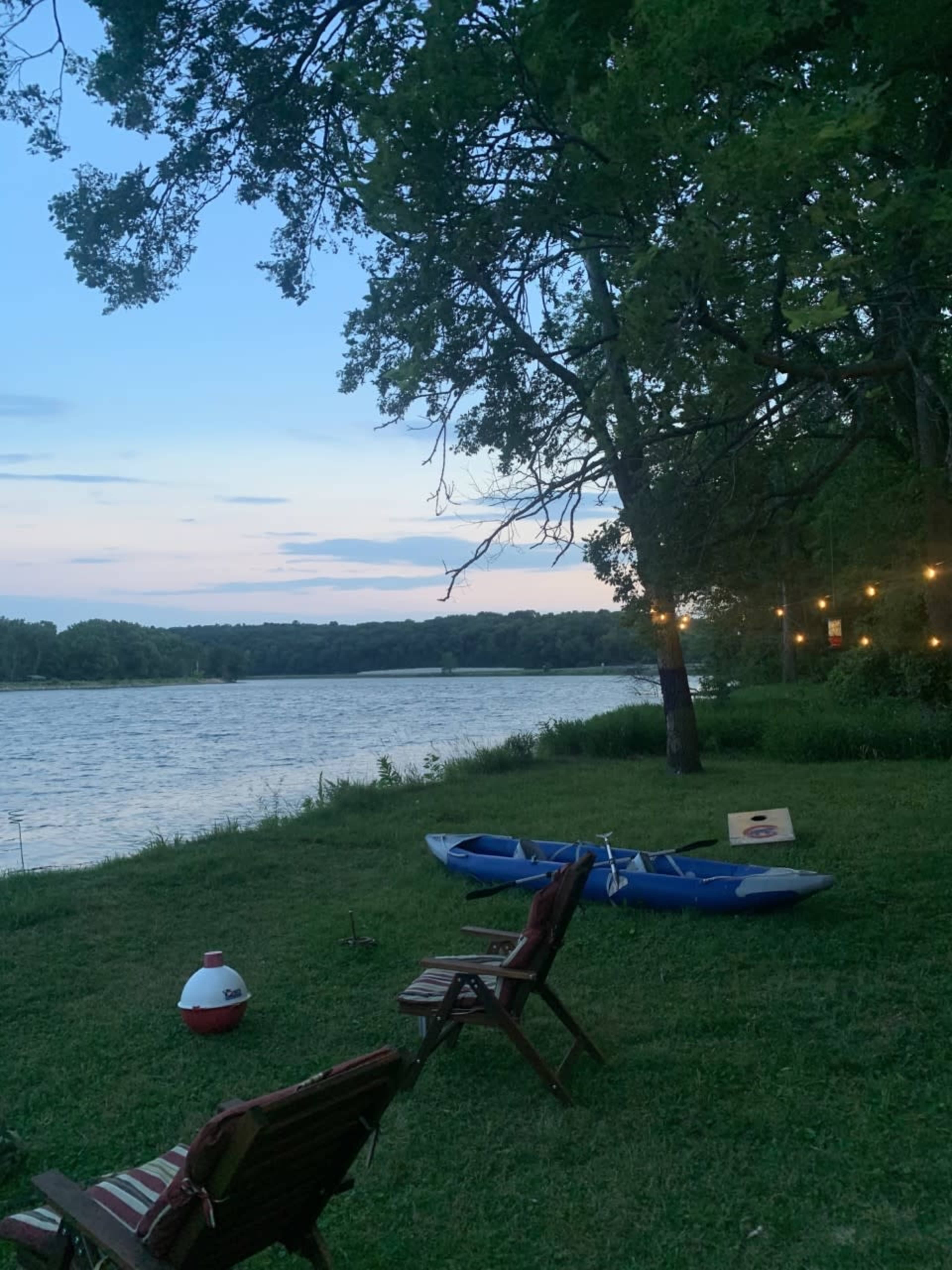 Two blue kayaks rest on the grass near a calm river under a twilight sky, with wooden chairs positioned nearby.