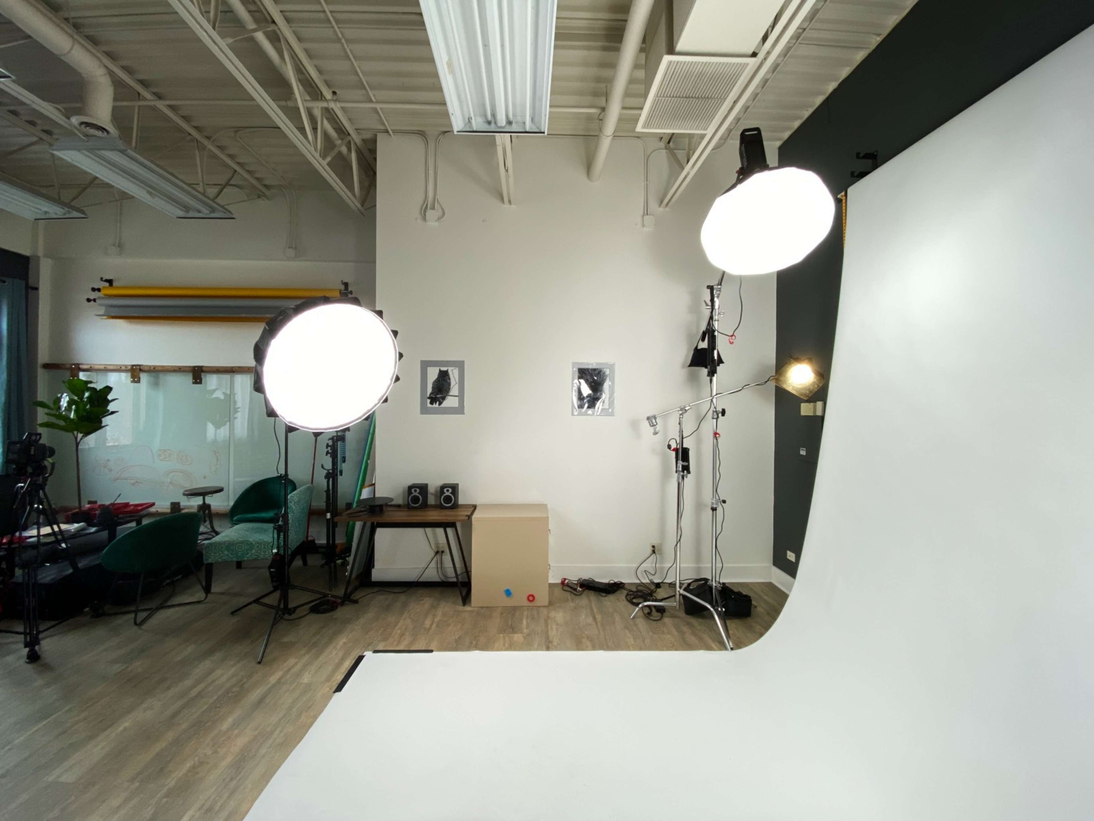 The image shows a photography studio with softboxes, a white backdrop, and a workspace with green chairs and framed black-and-white photographs.