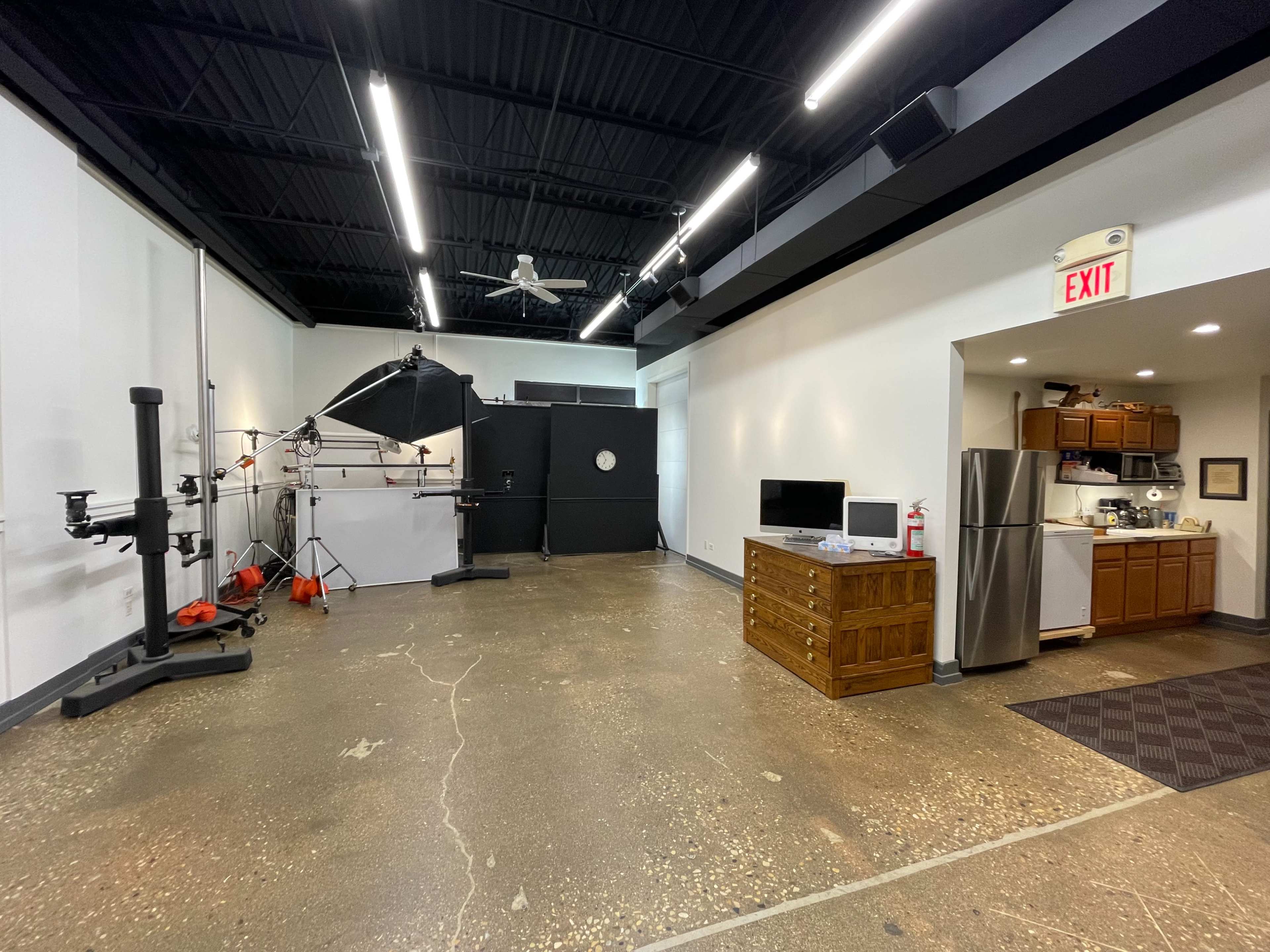 A spacious studio interior featuring a photography setup with lighting equipment, a counter with kitchen appliances, and an exit sign.
