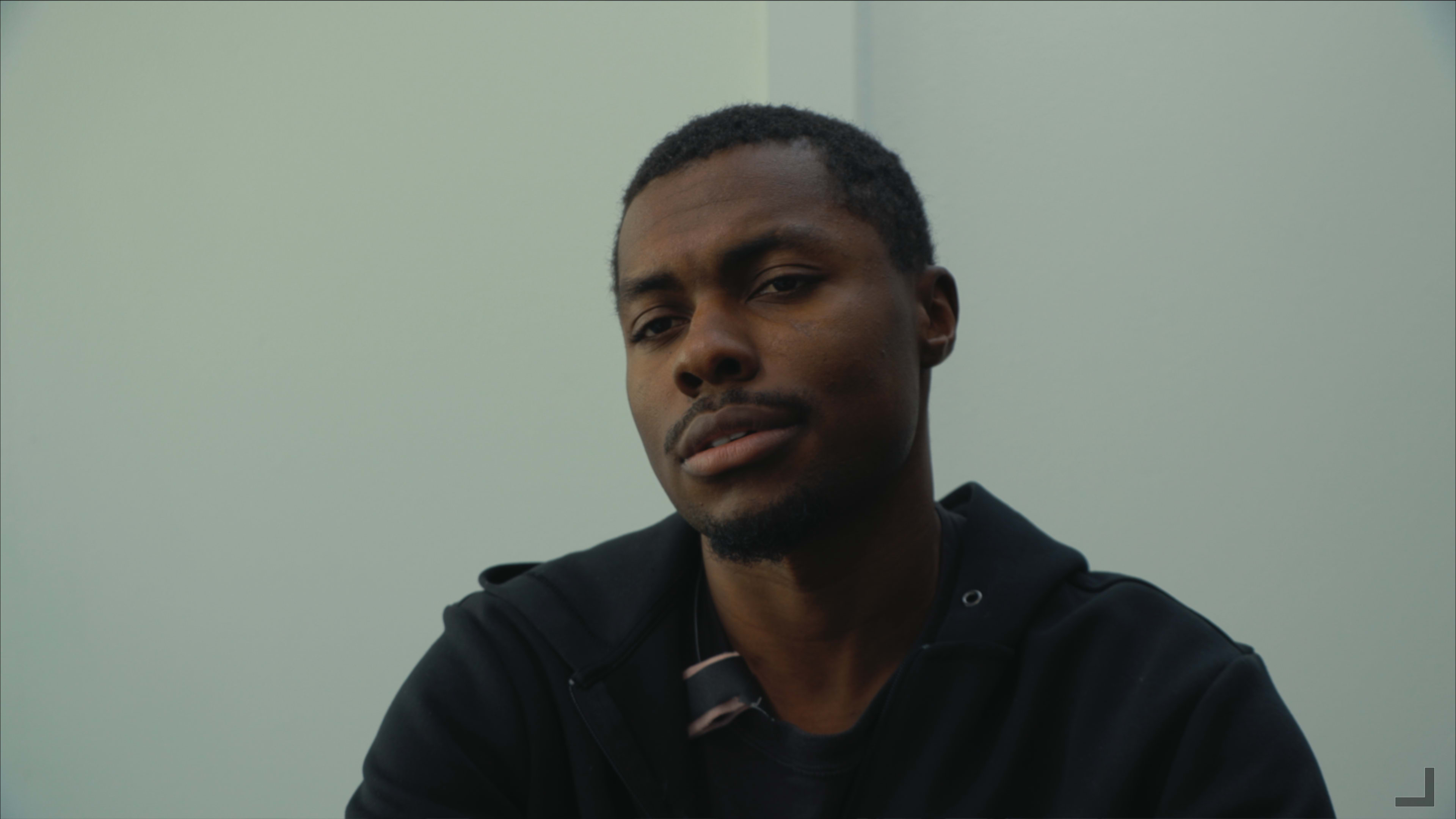 A man sits against a light-colored wall, looking directly at the camera with a neutral expression.