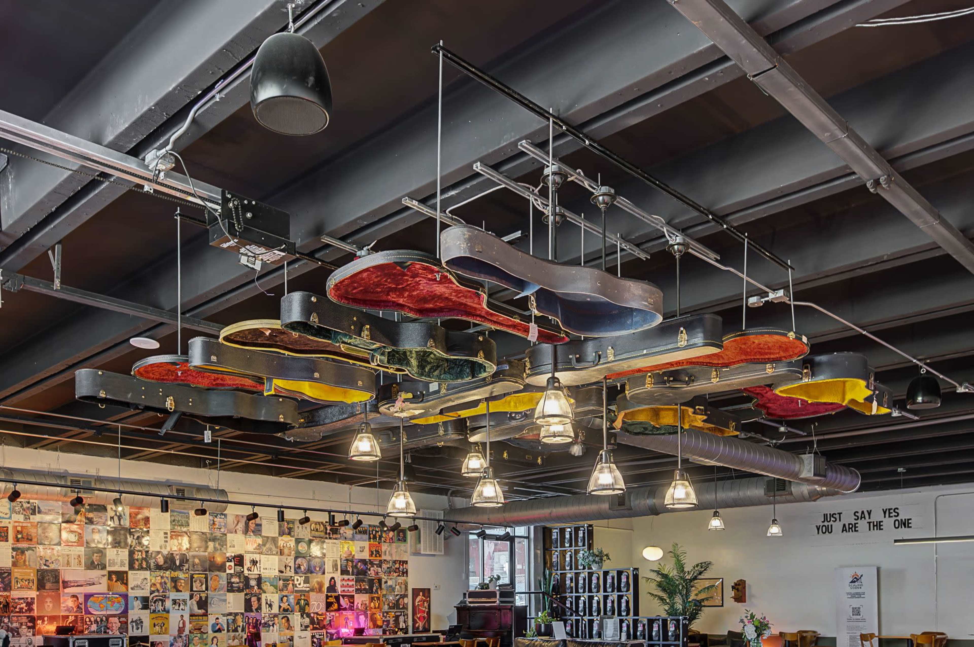 The image shows a ceiling installation featuring various guitar-shaped objects hanging above a space with pendant lights and a wall adorned with framed photos and records.