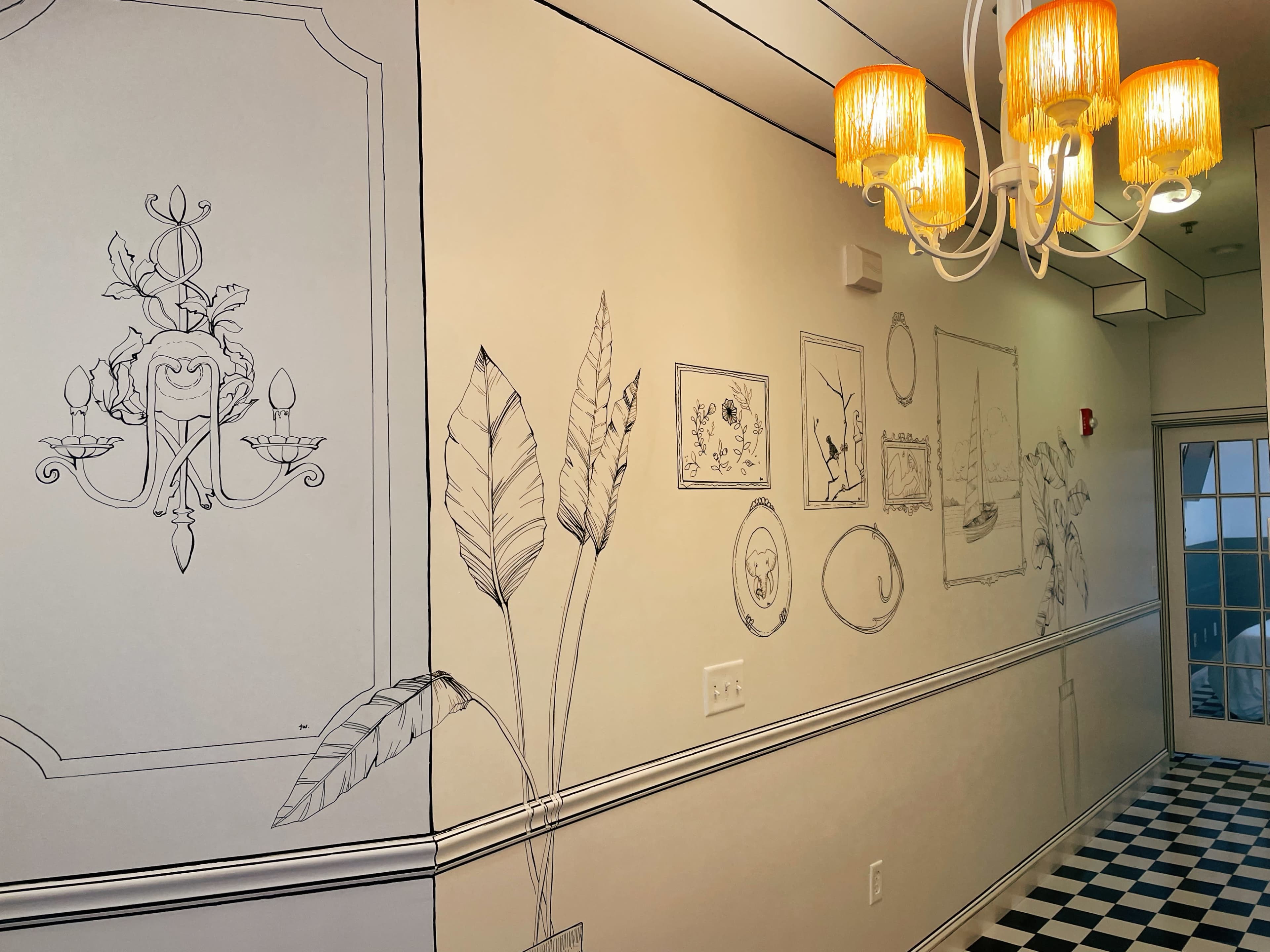 A hallway featuring hand-drawn wall decorations, including plants and framed artwork, with a chandelier providing overhead light.