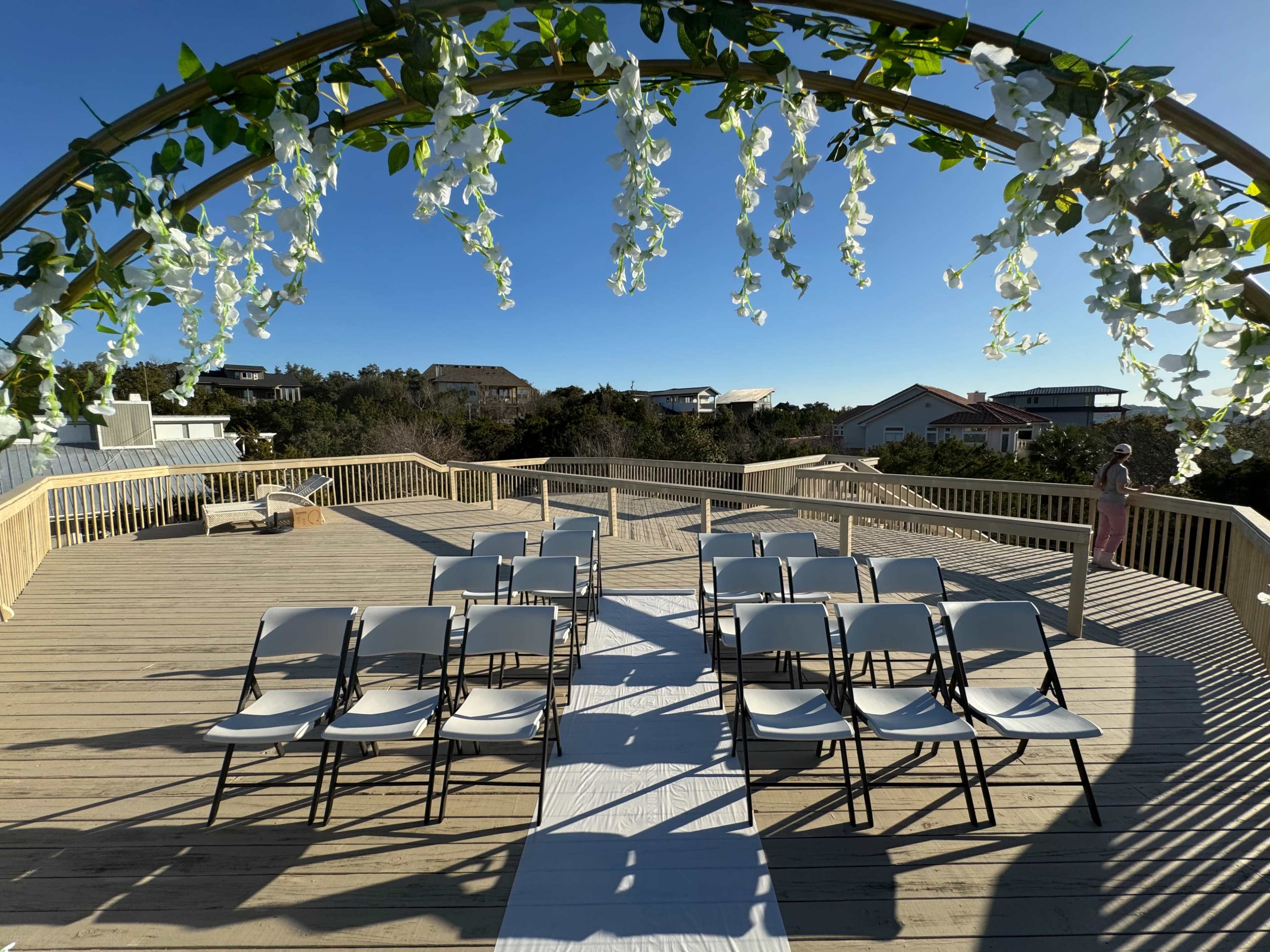 A wooden deck is set up for a wedding ceremony, featuring rows of white chairs and a floral arch, with a clear blue sky above.