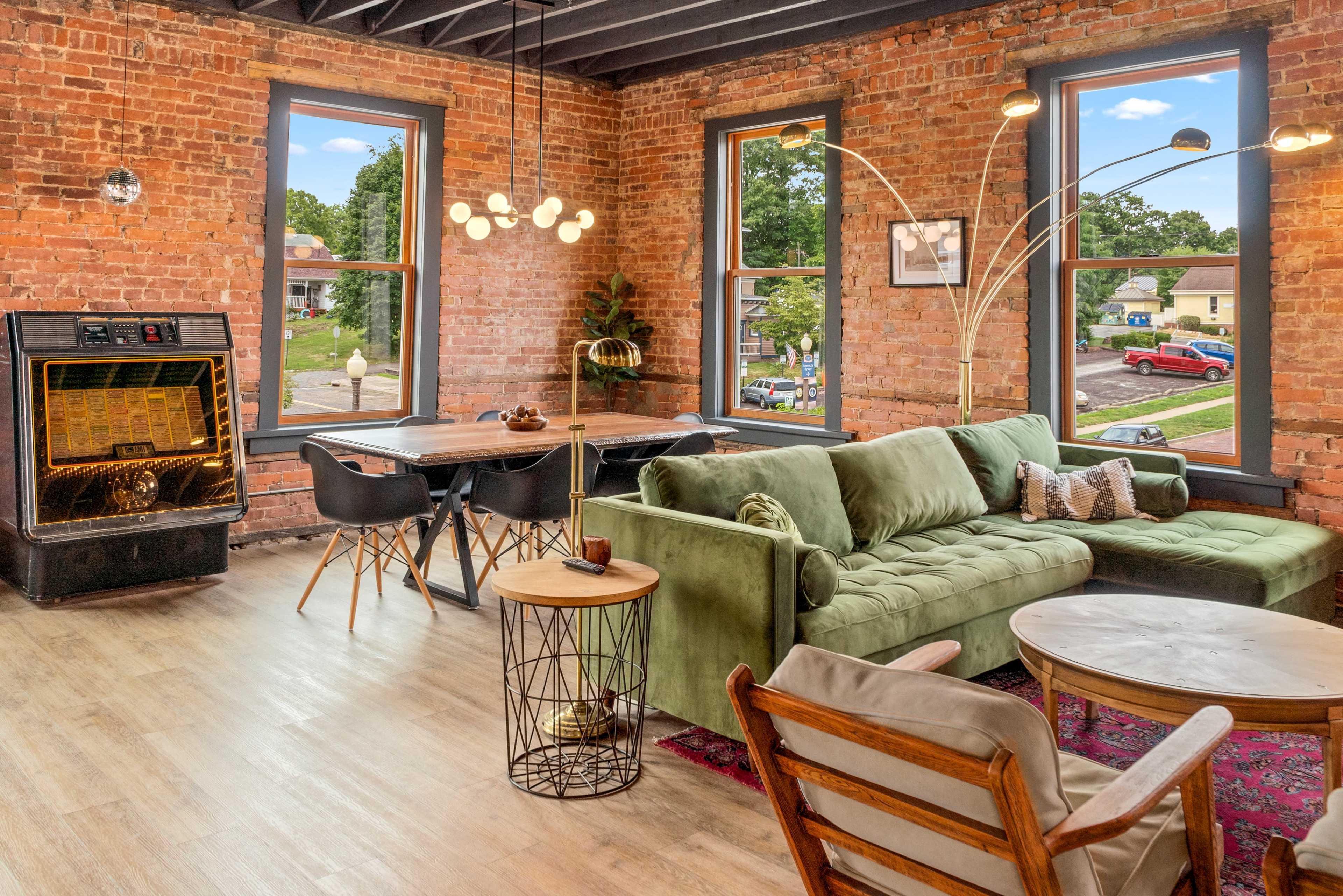 A cozy interior with exposed brick walls, a green sectional sofa, a dining area, and large windows allowing natural light to flood the space.