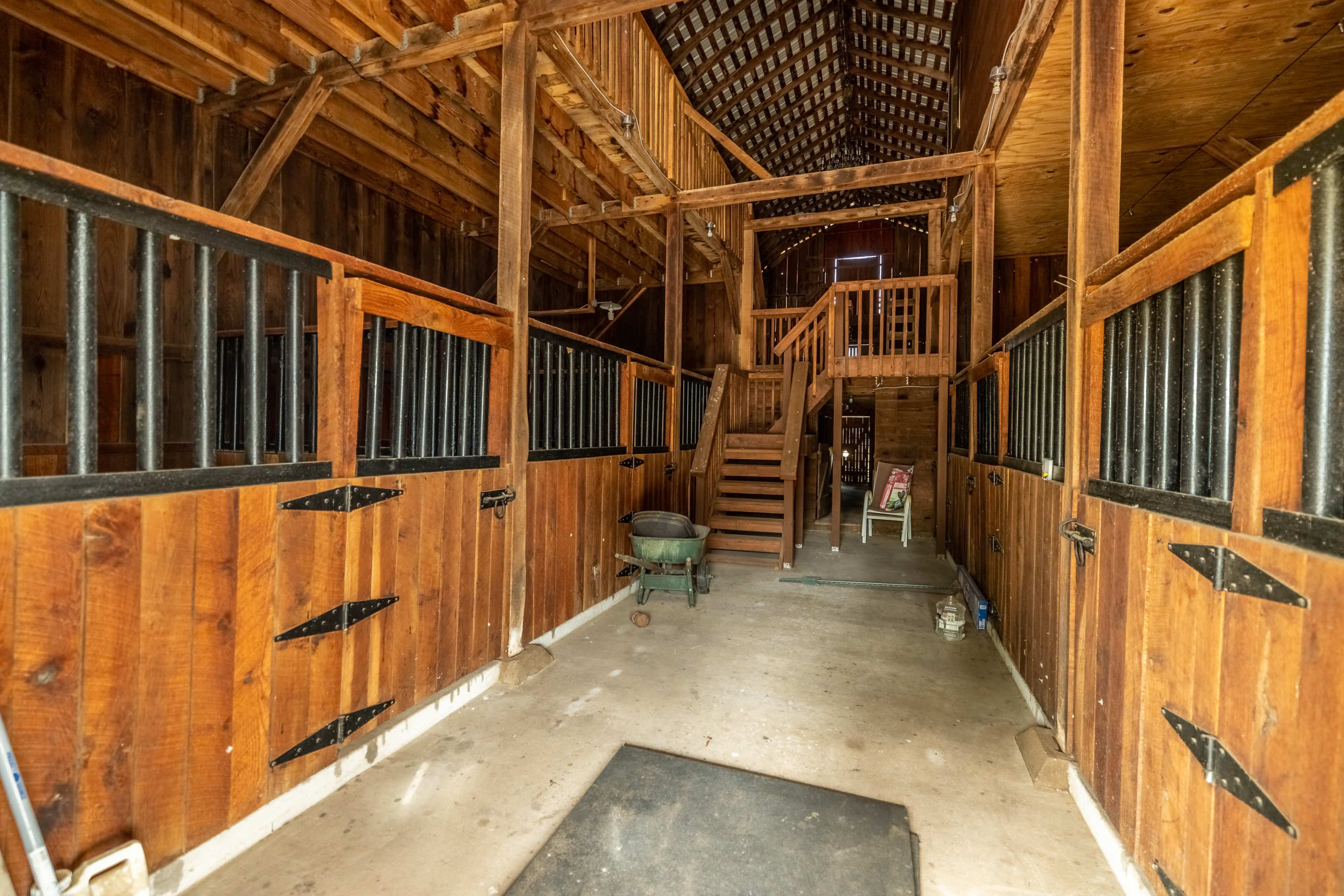 The interior of a rustic barn with wooden stalls, a staircase leading to an upper level, and a concrete floor.