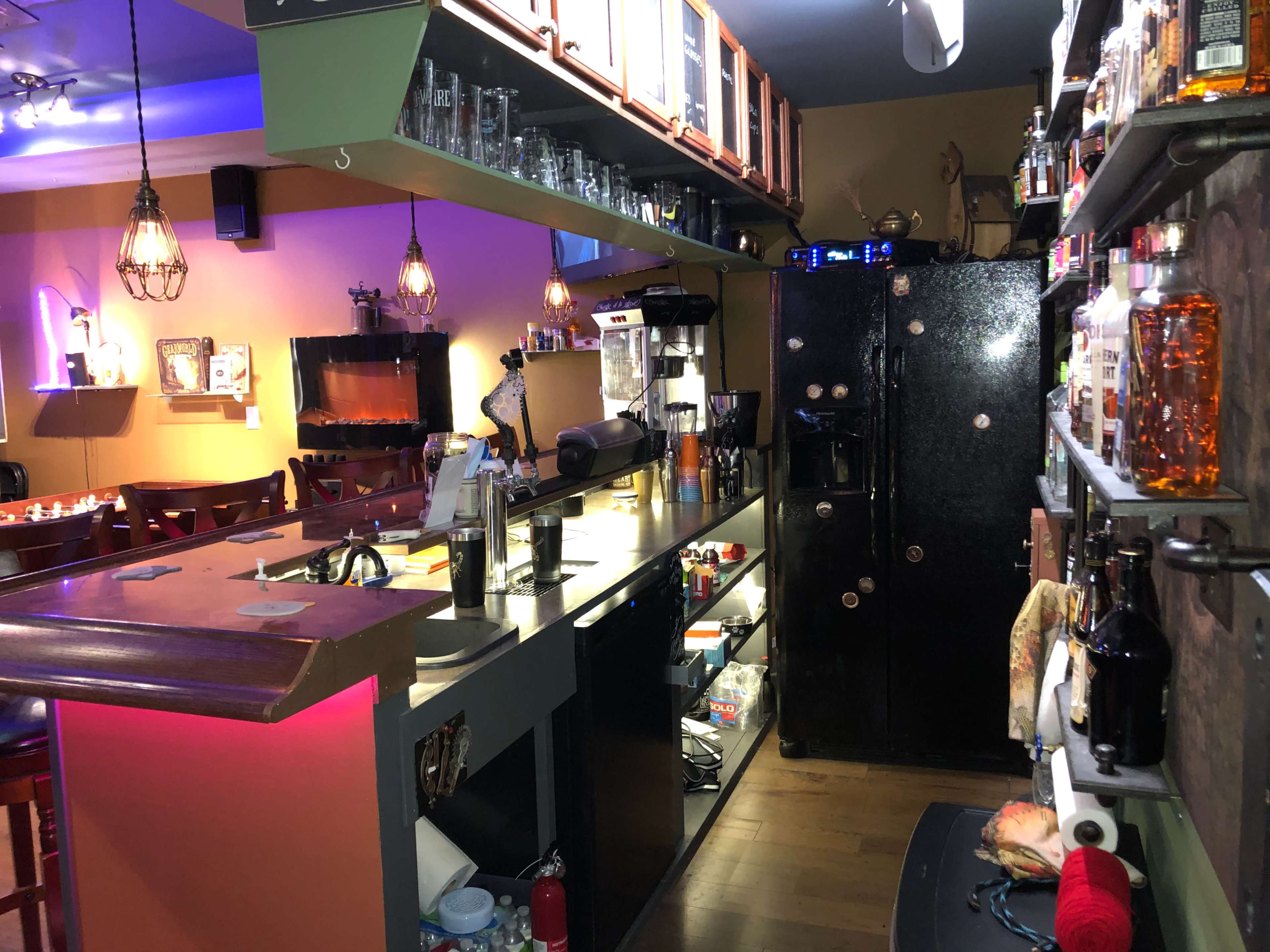 A cozy bar area with a counter, shelves lined with bottles, and a black refrigerator in a warmly lit room.