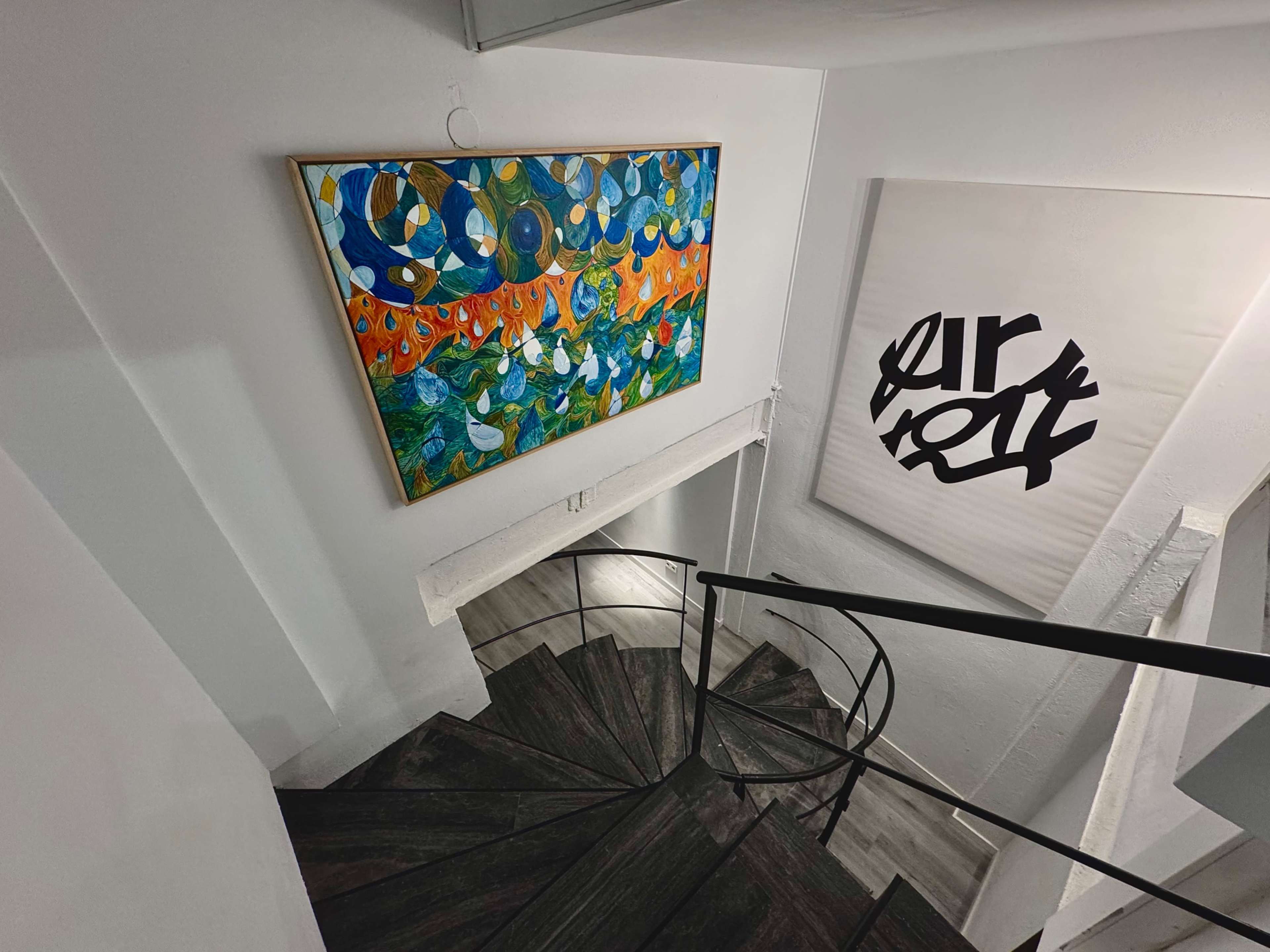 A colorful painting hangs on a wall above a spiral staircase in a brightly lit interior space.