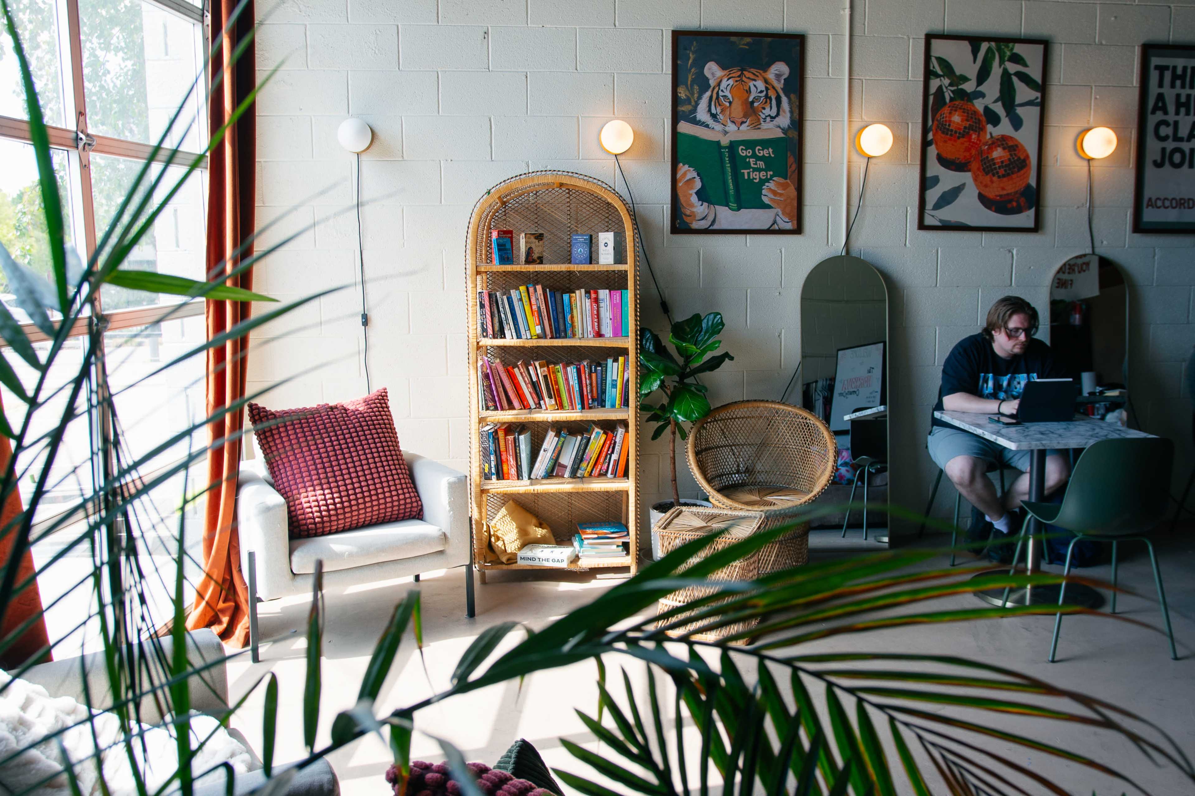 The image shows a cozy indoor space with a bookshelf, seating areas, and a person working on a laptop at a table, surrounded by decorative plants and artwork on the walls.