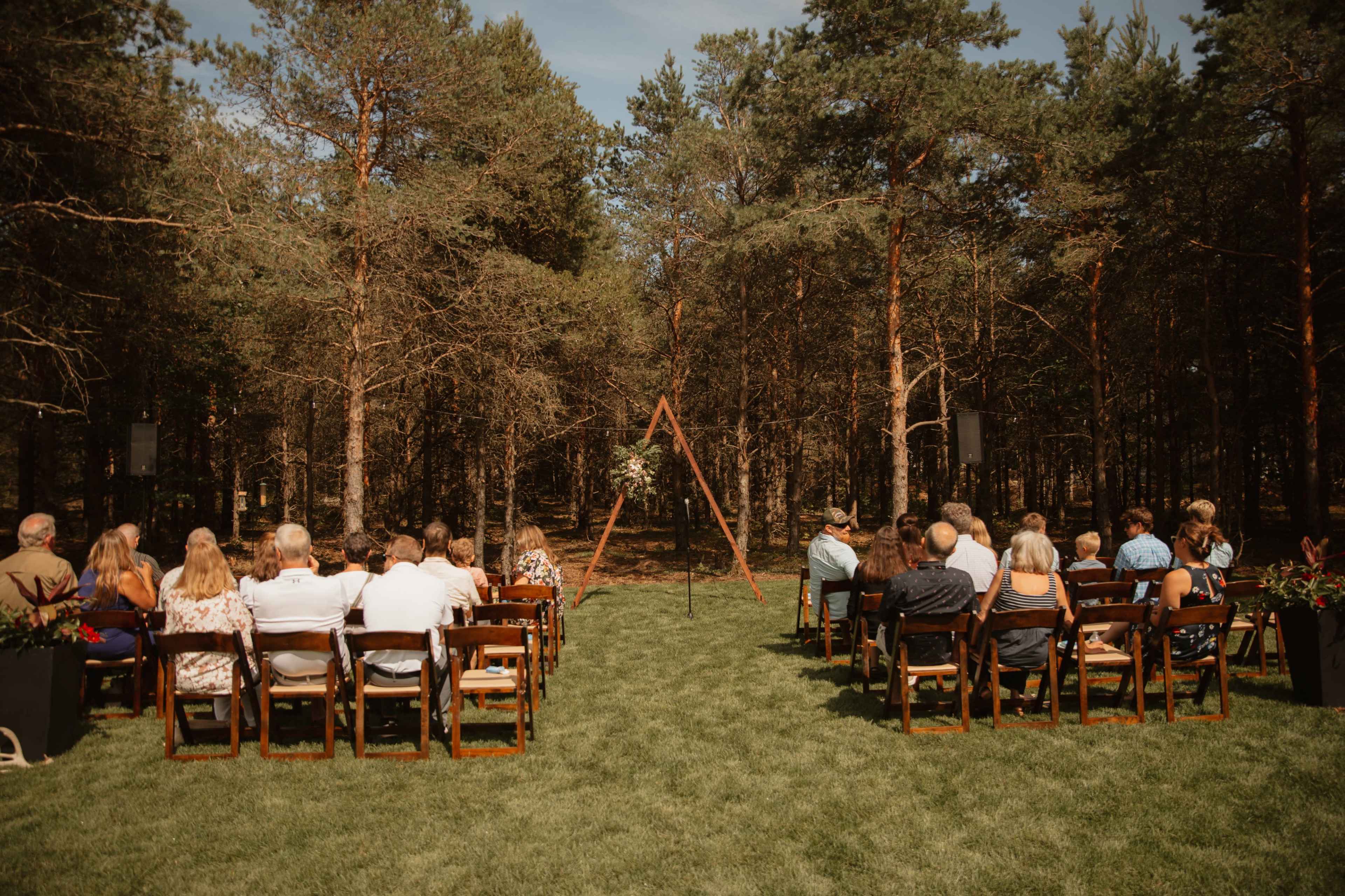 A wedding ceremony is set up in a forest clearing with guests seated on wooden chairs facing a floral-decorated arch.