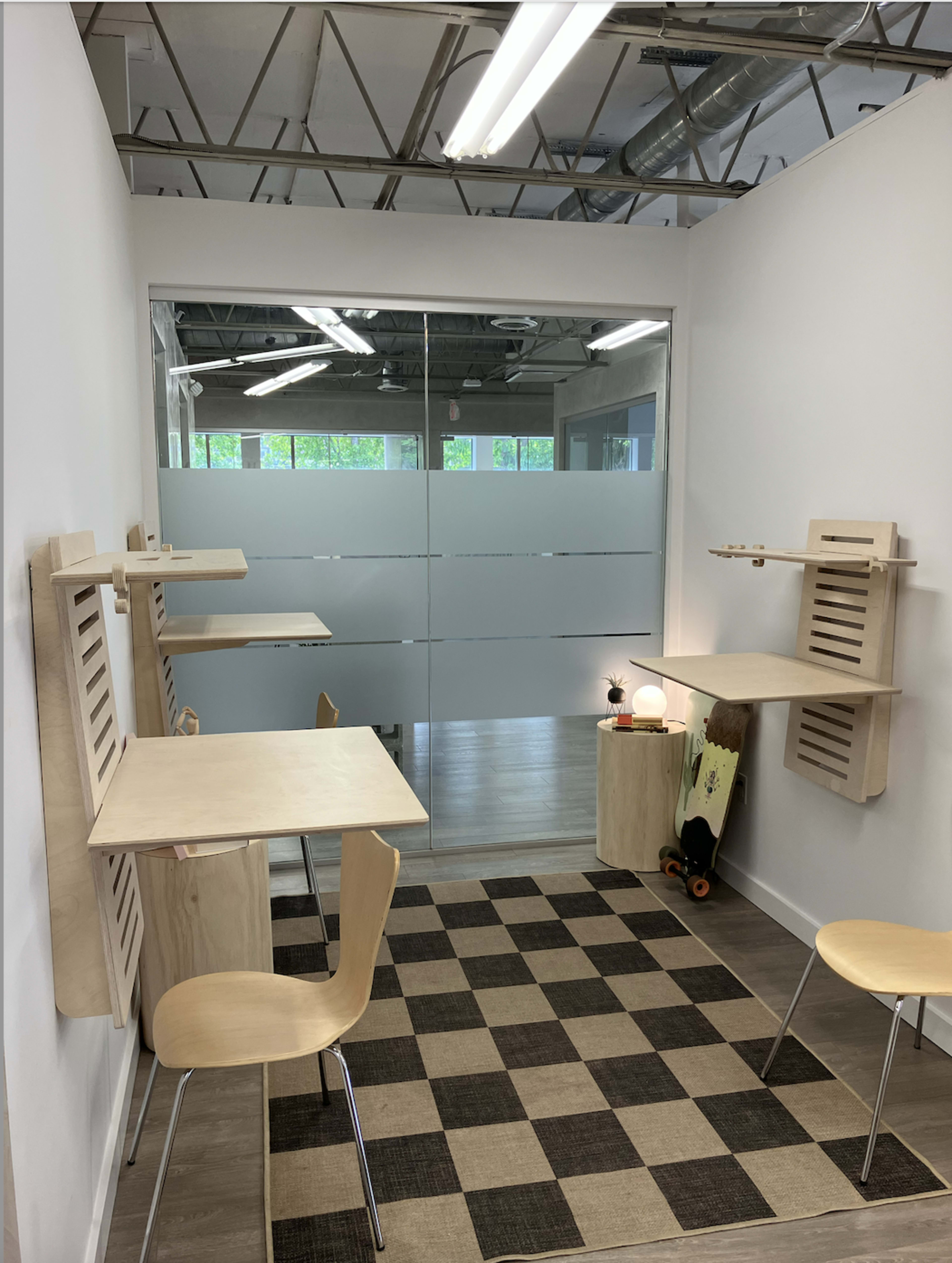 The image shows a minimalist workspace with two wooden desks attached to the walls, each accompanied by a chair, and a checkered rug on the floor.