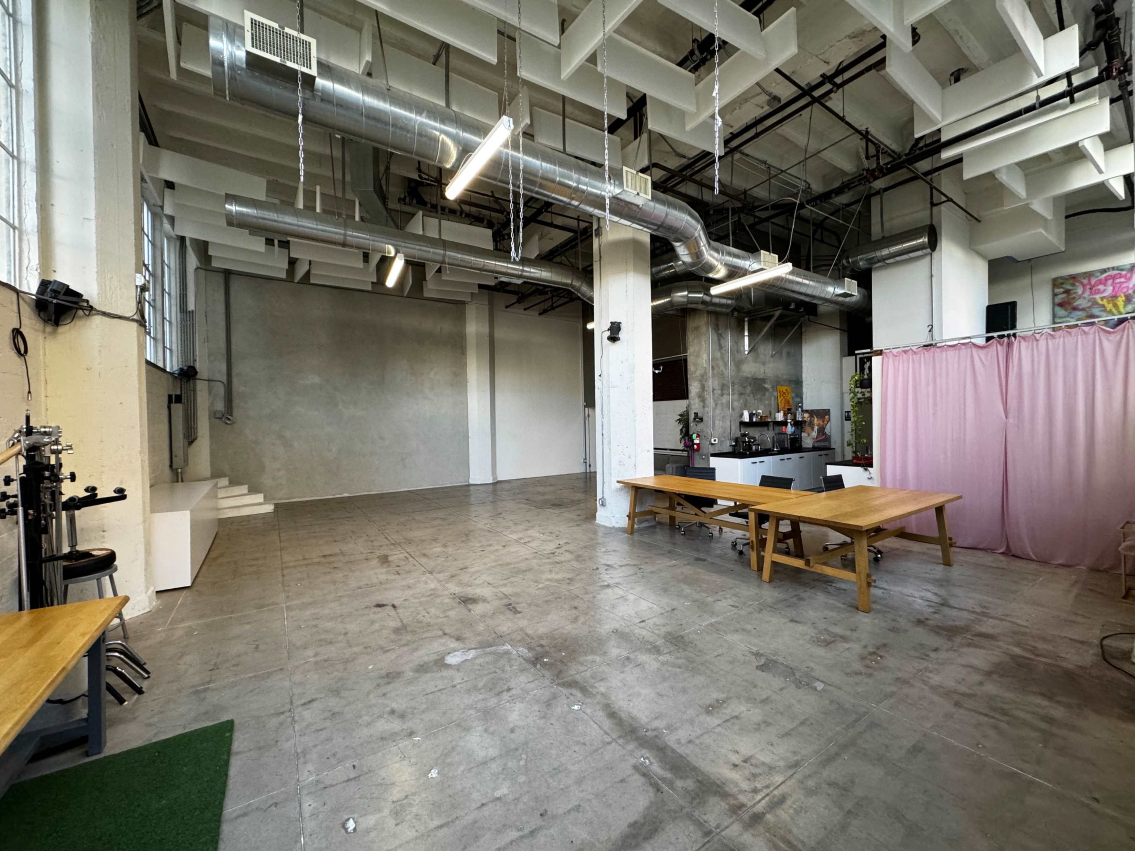 A spacious, industrial-style room with concrete floors, large windows, and a pink curtain separating an area with a kitchen and dining table.