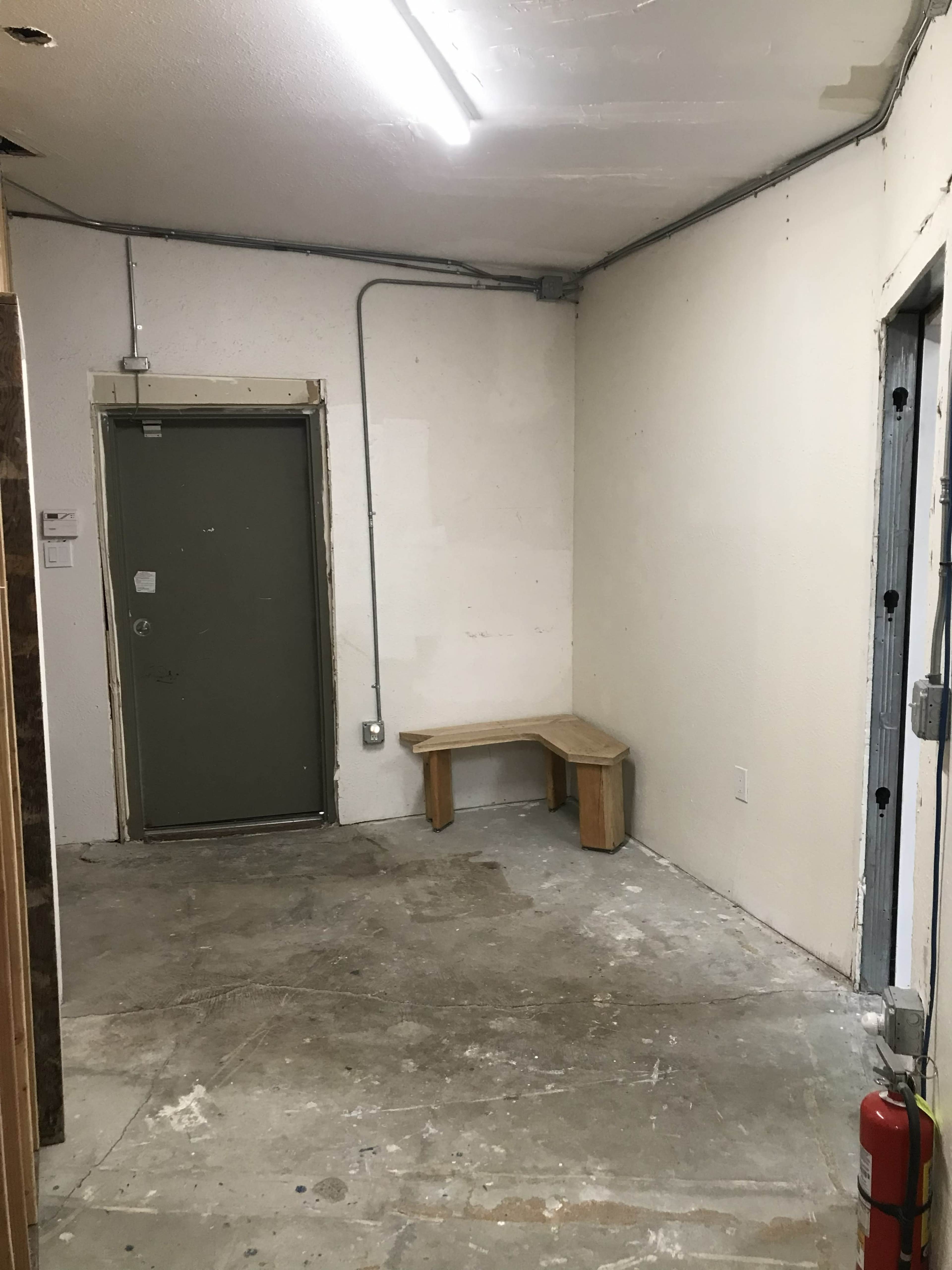 The image shows a small, empty room with a wooden bench in one corner and a door on the adjacent wall.