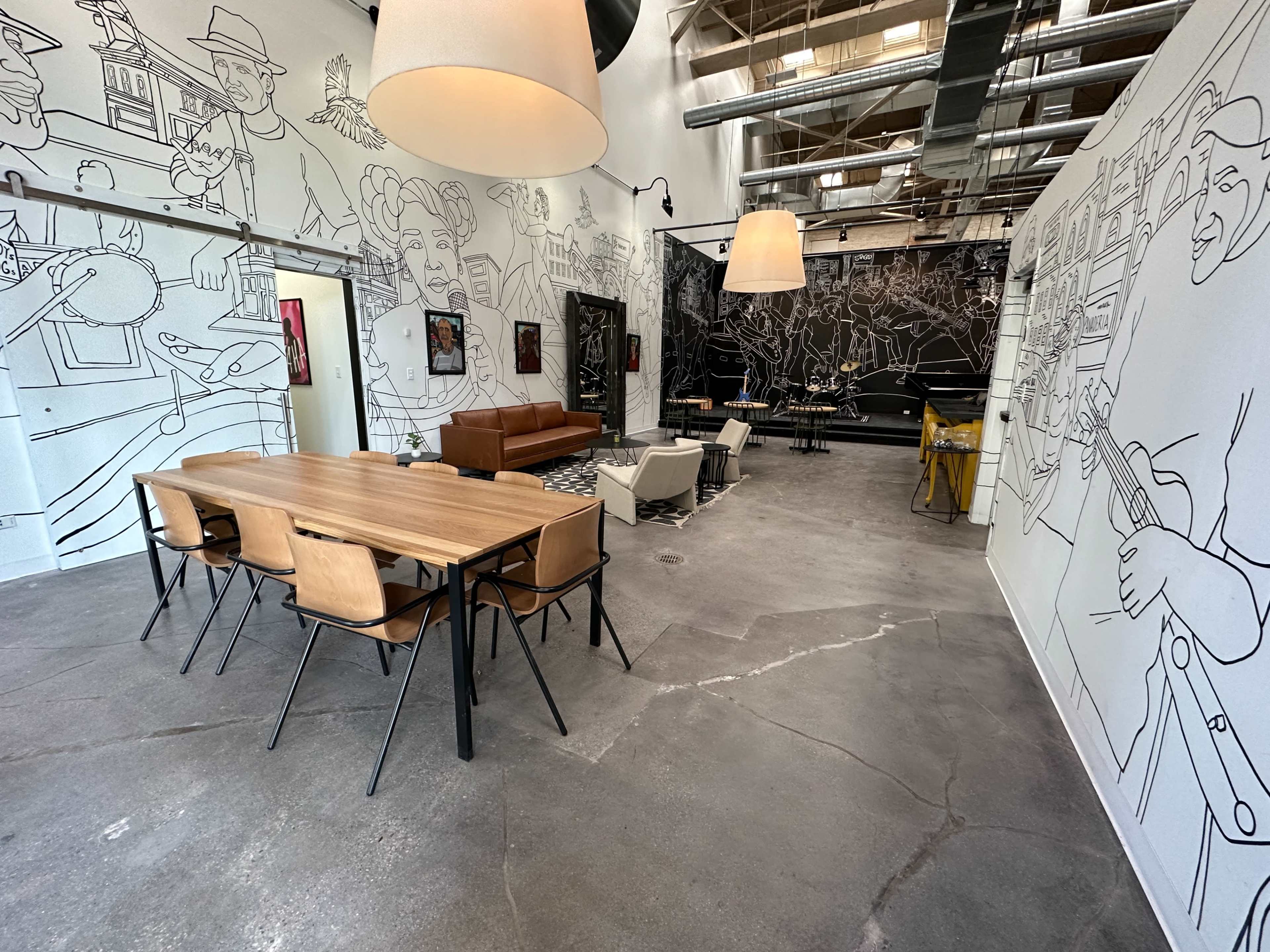 An industrial-style interior with a dining table, chairs, a brown sofa, and large murals on the walls.