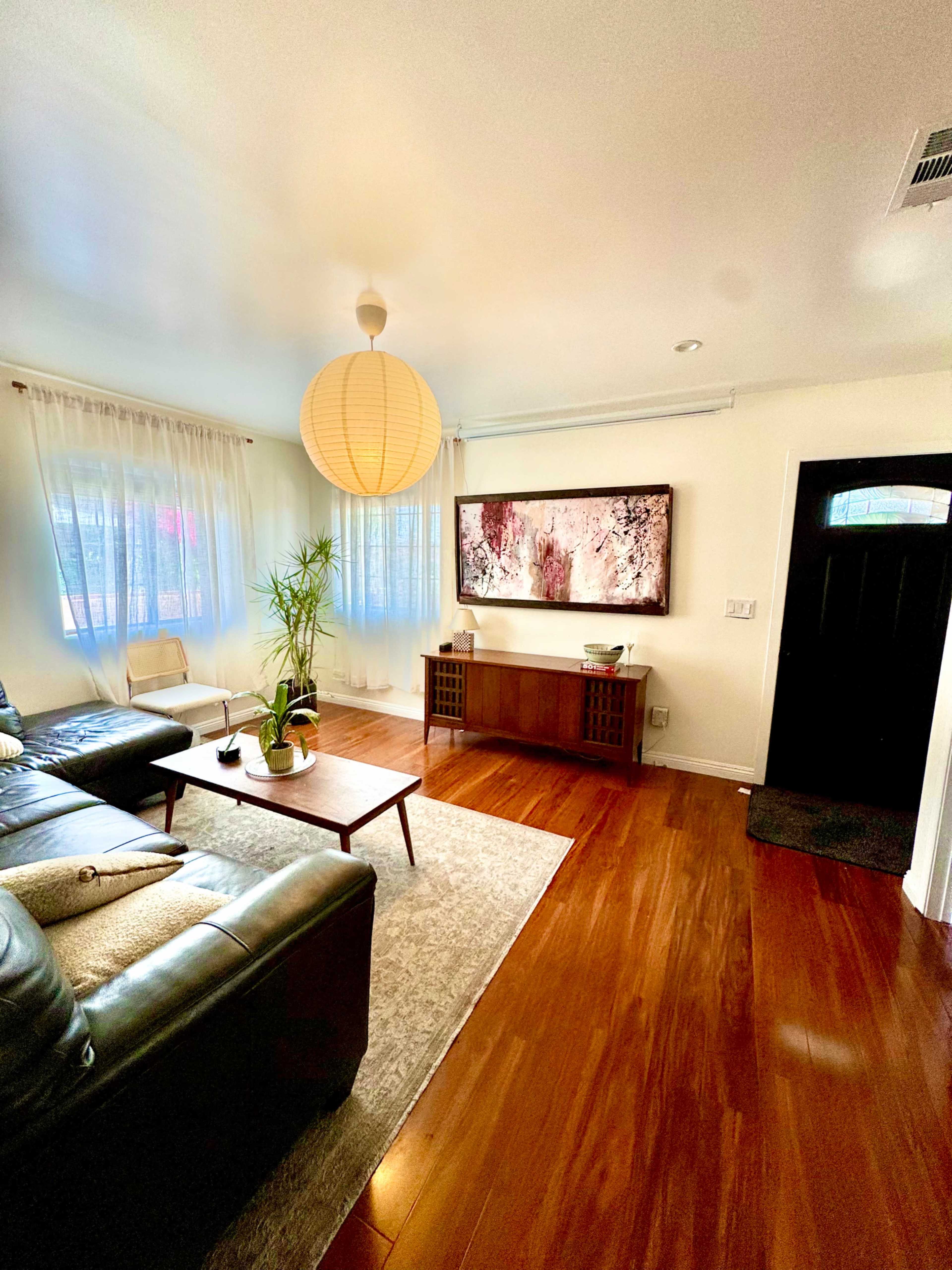 The image shows a well-lit living room with hardwood floors, a leather sofa, a coffee table, and a large wall-mounted artwork, along with a front door visible on the right.