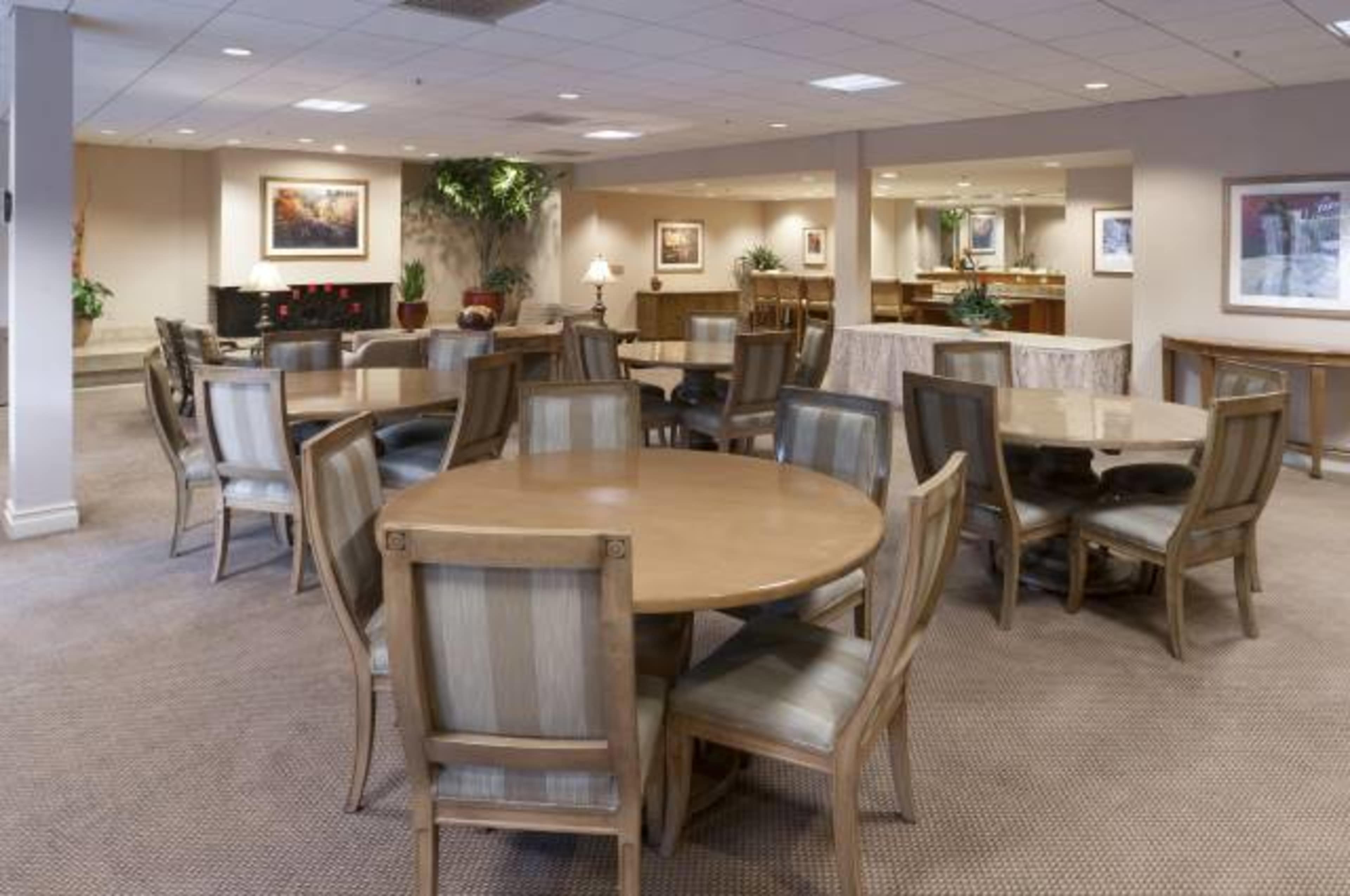 A spacious dining area with several round wooden tables and chairs arranged on a carpeted floor, surrounded by plants and framed artwork.