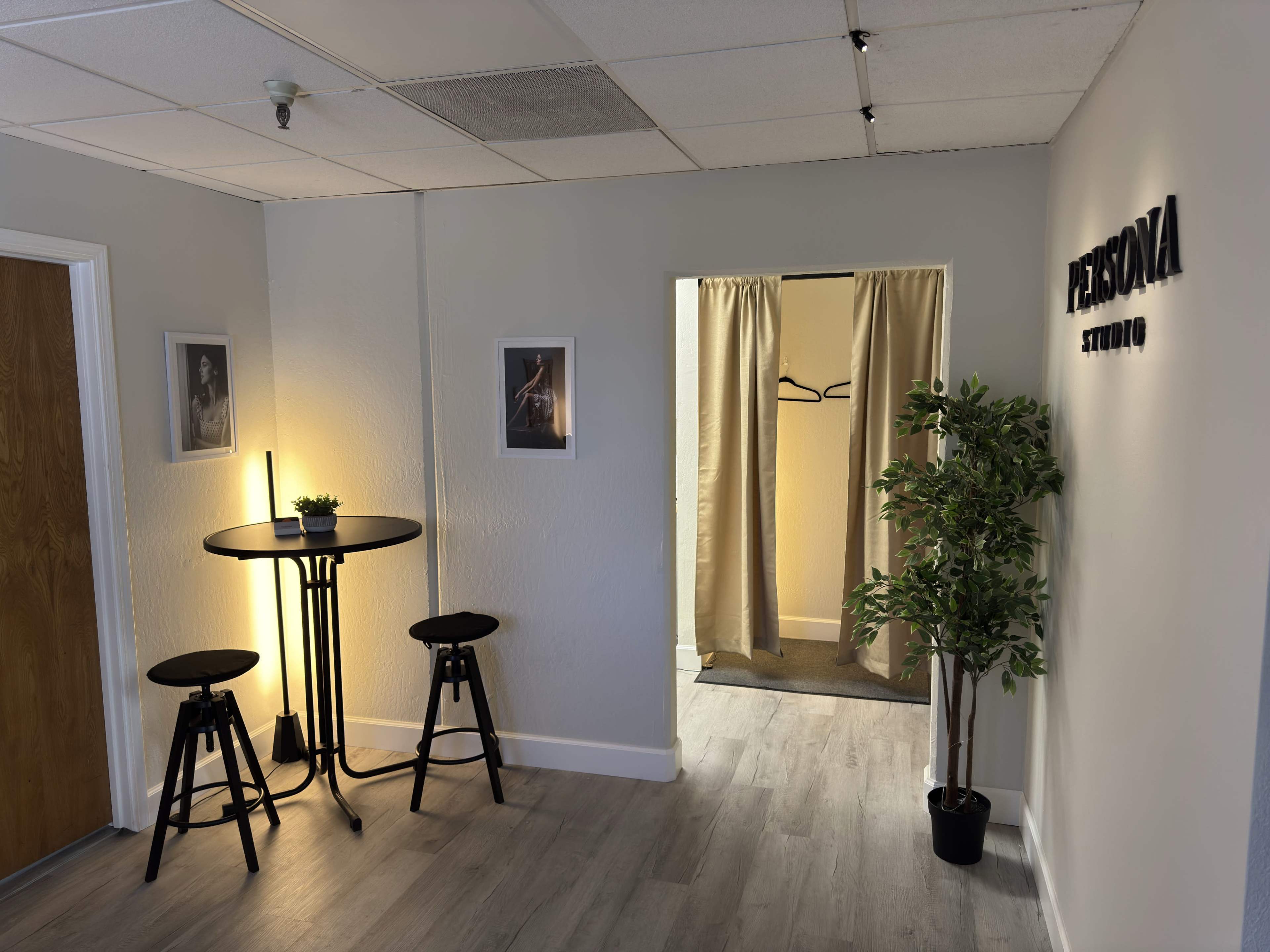 The image shows a small waiting area with two bar stools, a round table, a small plant, and a doorway draped with curtains leading to another room.