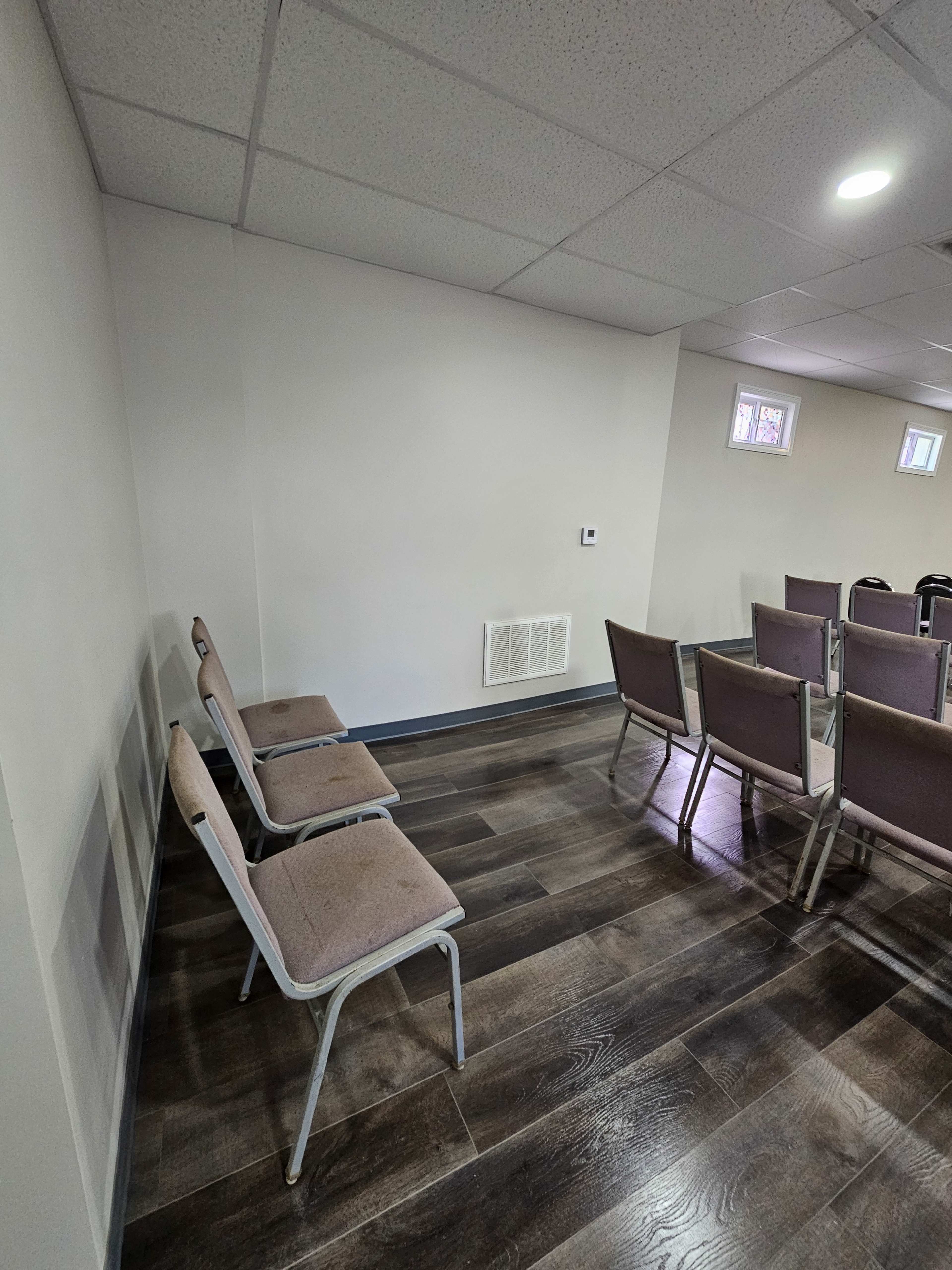 The image shows a room with several stacked and unstacked chairs against a wall, featuring a wooden floor and plain walls.