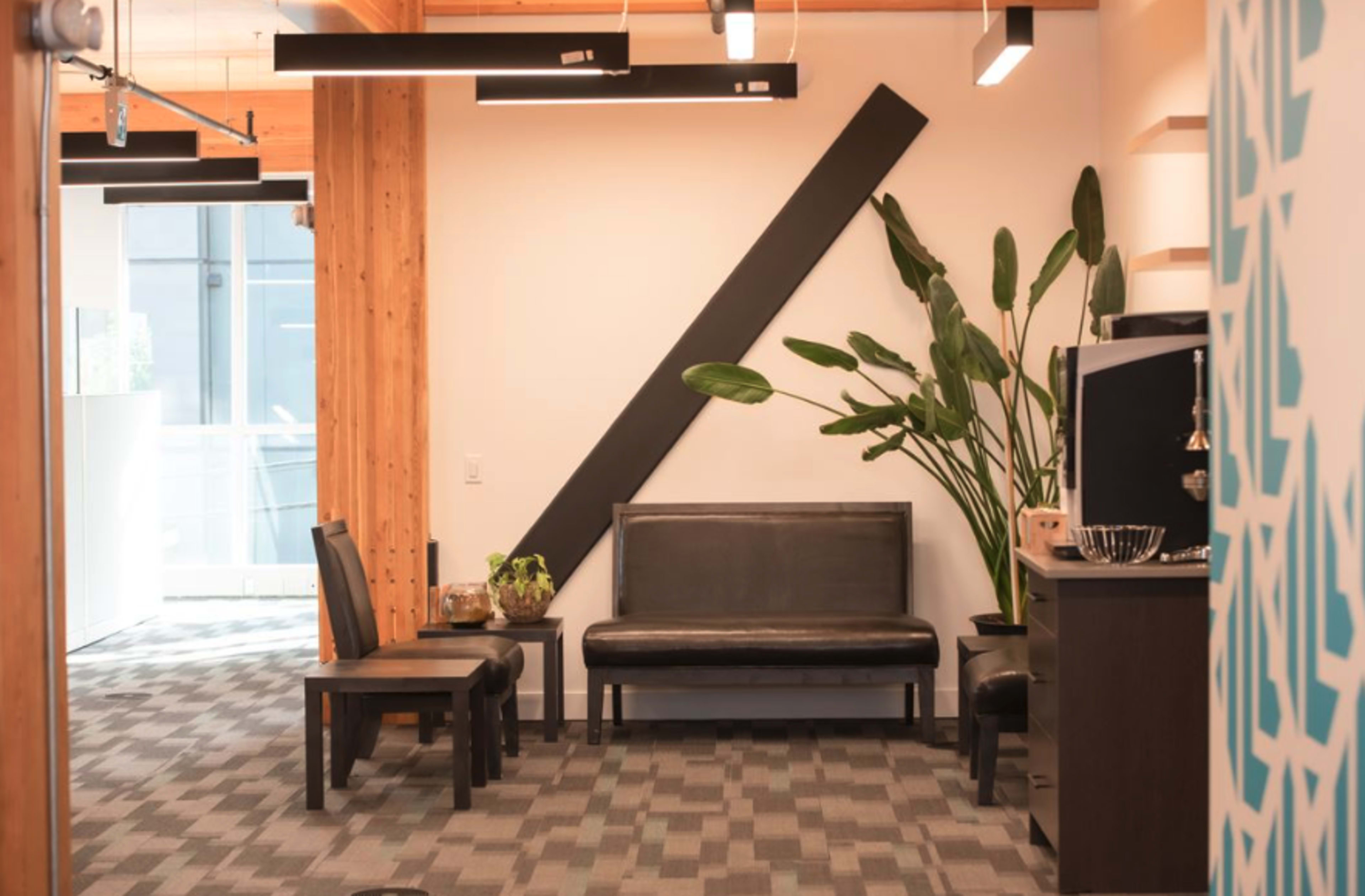 A modern office waiting area with a black bench, two chairs, a potted plant, and a dark cabinet against a light wall.