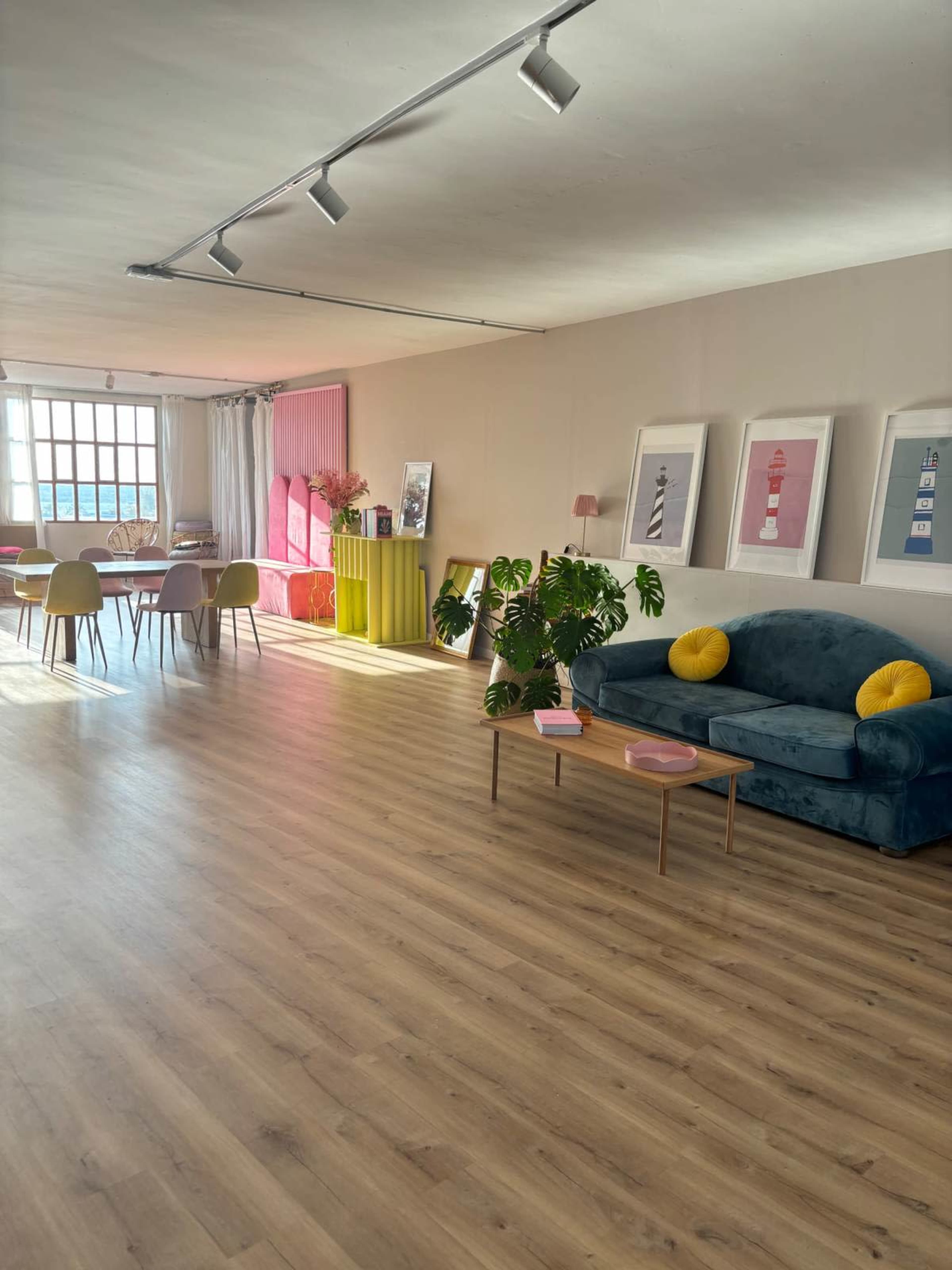 The image shows a bright, modern interior space with a blue couch, a yellow accent table, a dining area, and colorful decor on the walls.
