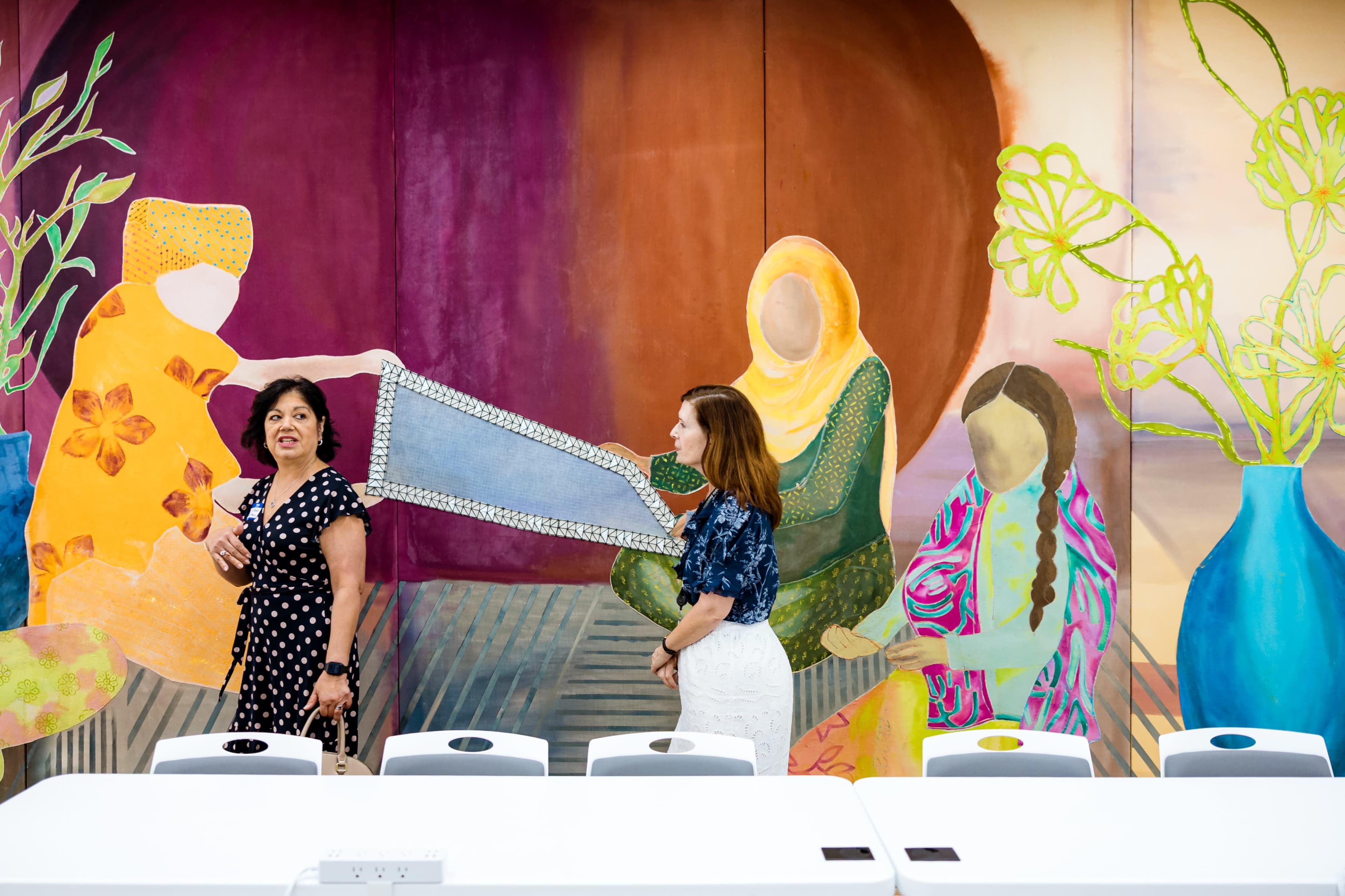 Two women are engaged in conversation in front of a vibrant mural depicting colorful figures and plants in a meeting space.