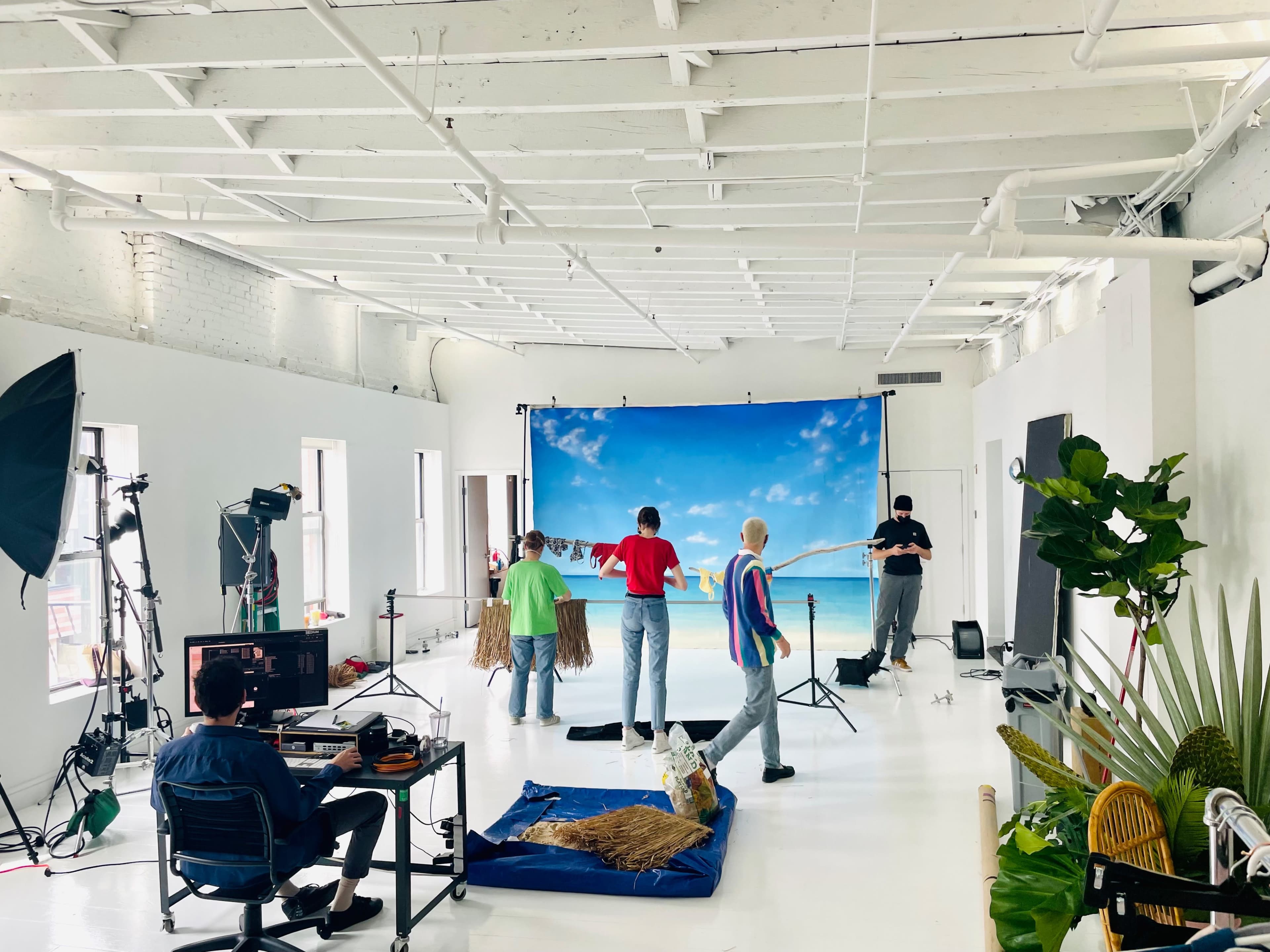 A film studio setup is shown with a blue backdrop depicting a beach scene, while several people and equipment are arranged for a photoshoot.
