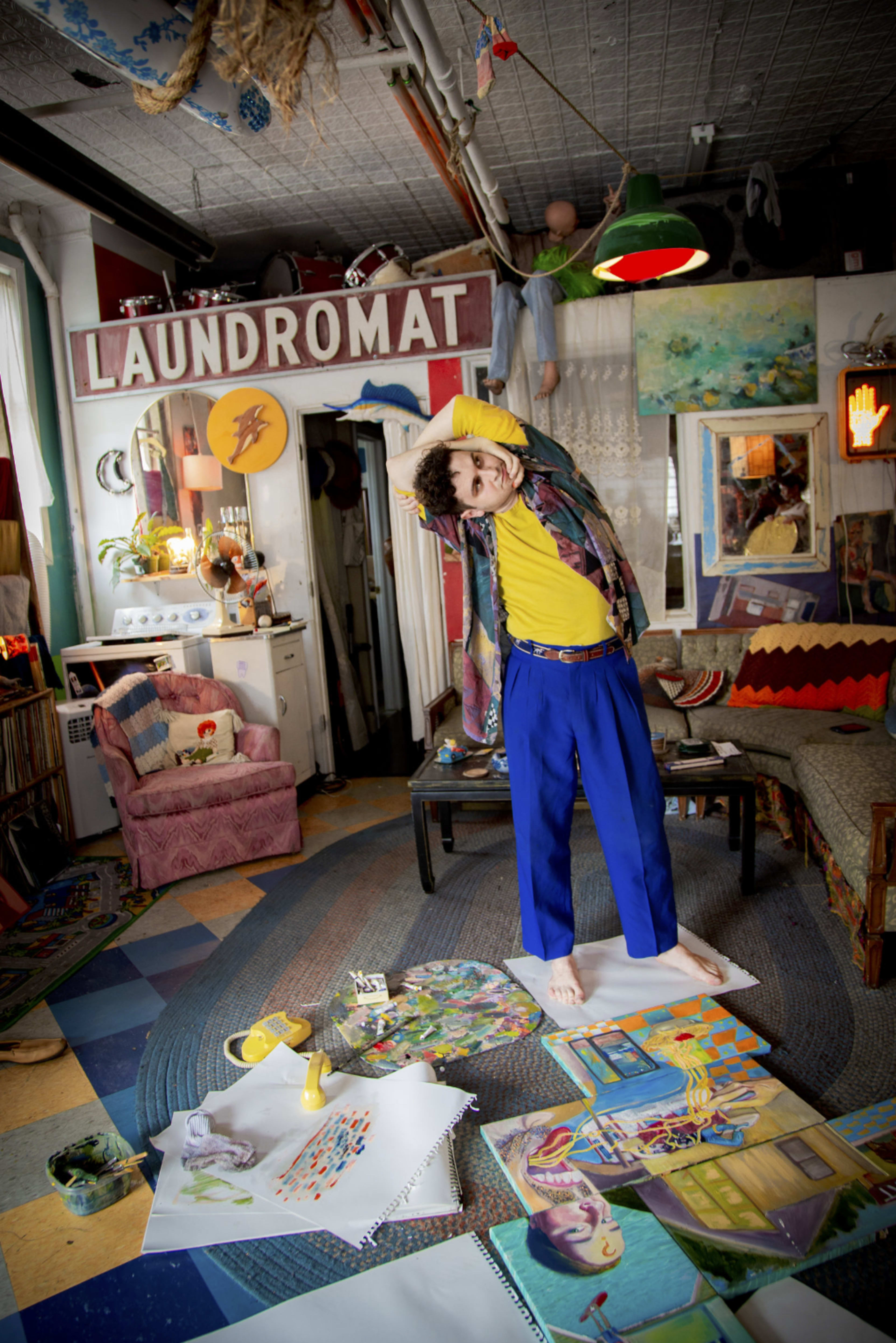 A person in a colorful outfit stands on a floor covered with various art supplies and paintings inside a creatively decorated room with a "LAUNDROMAT" sign.