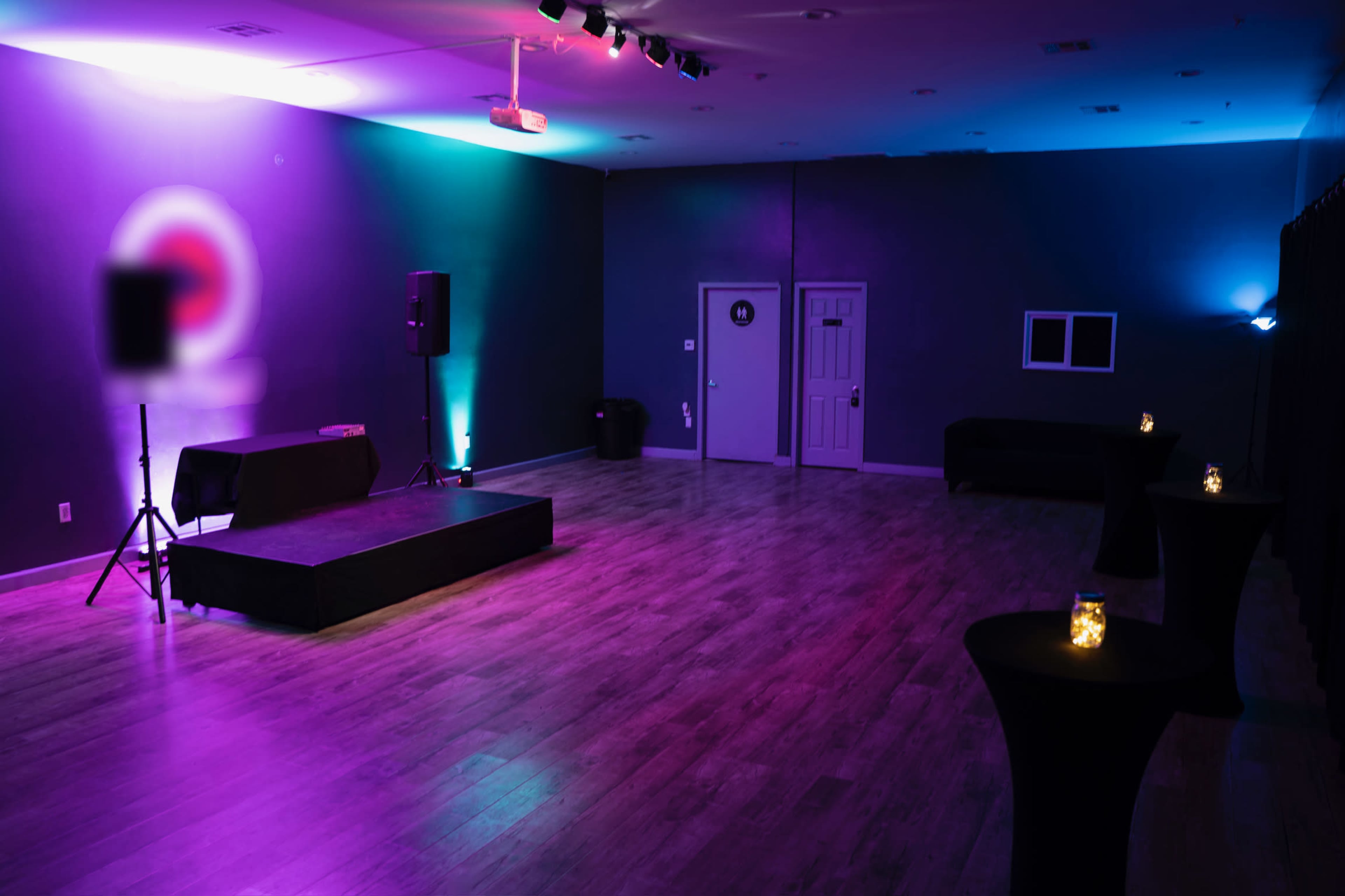 A dimly lit event space with purple and teal lighting, featuring a small stage, a sound speaker, and several candle-lit tables.