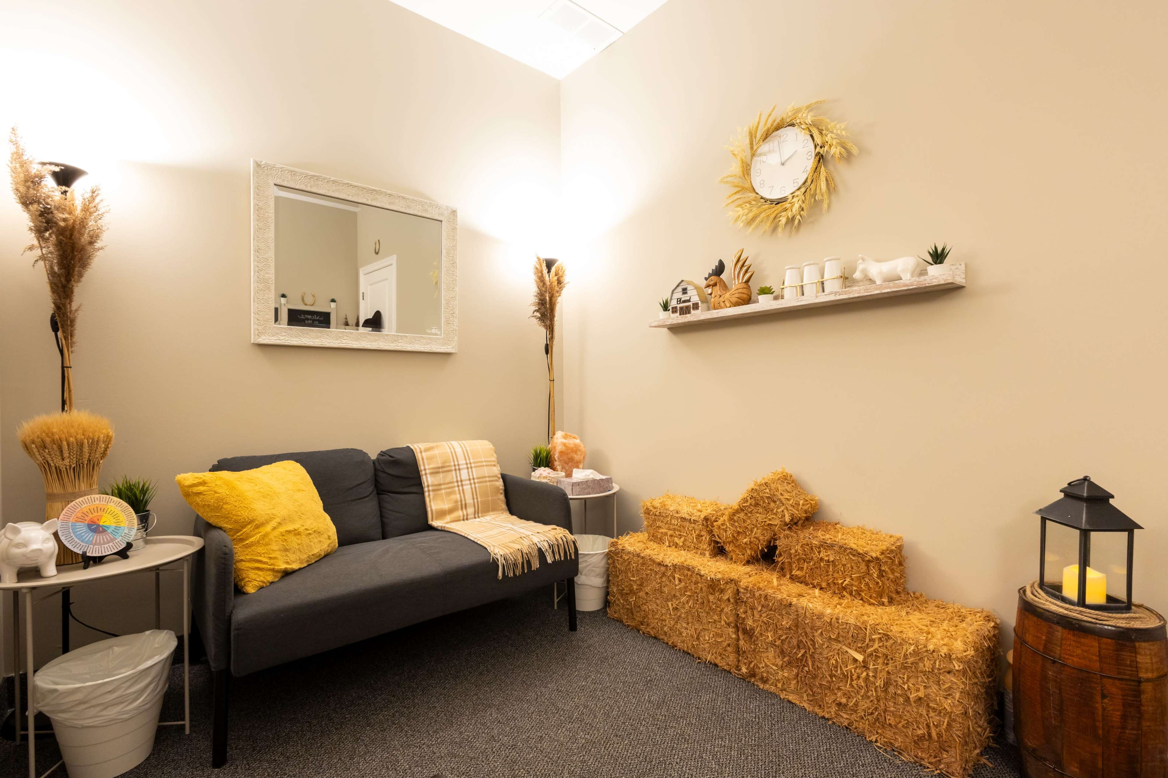 The scene depicts a cozy room with a gray sofa, decorative straw bales, a mirror, and a wall shelf displaying various items.
