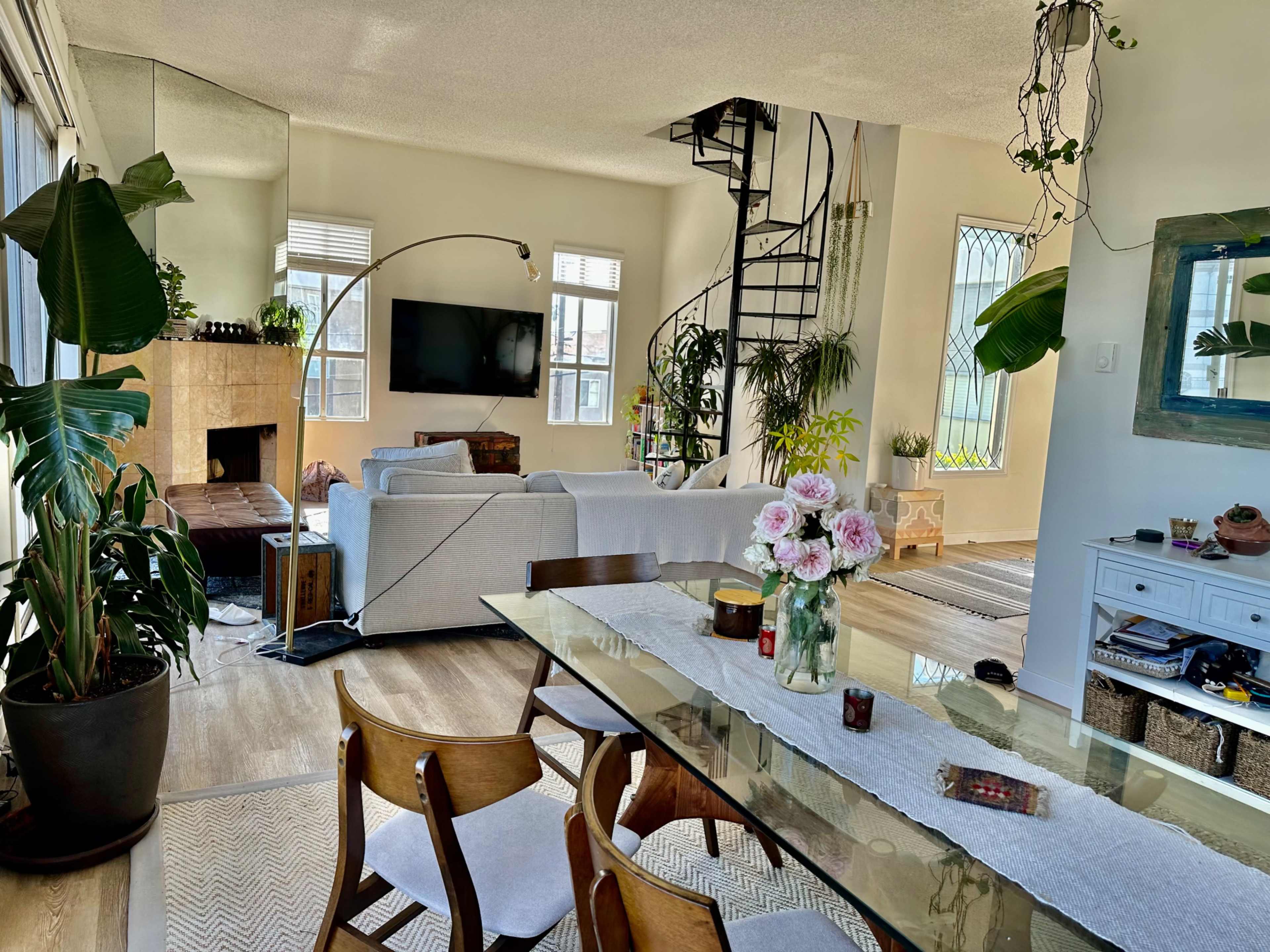 A bright, open living space with a spiral staircase, a large sectional sofa, a dining table set with flowers, and various indoor plants throughout the room.