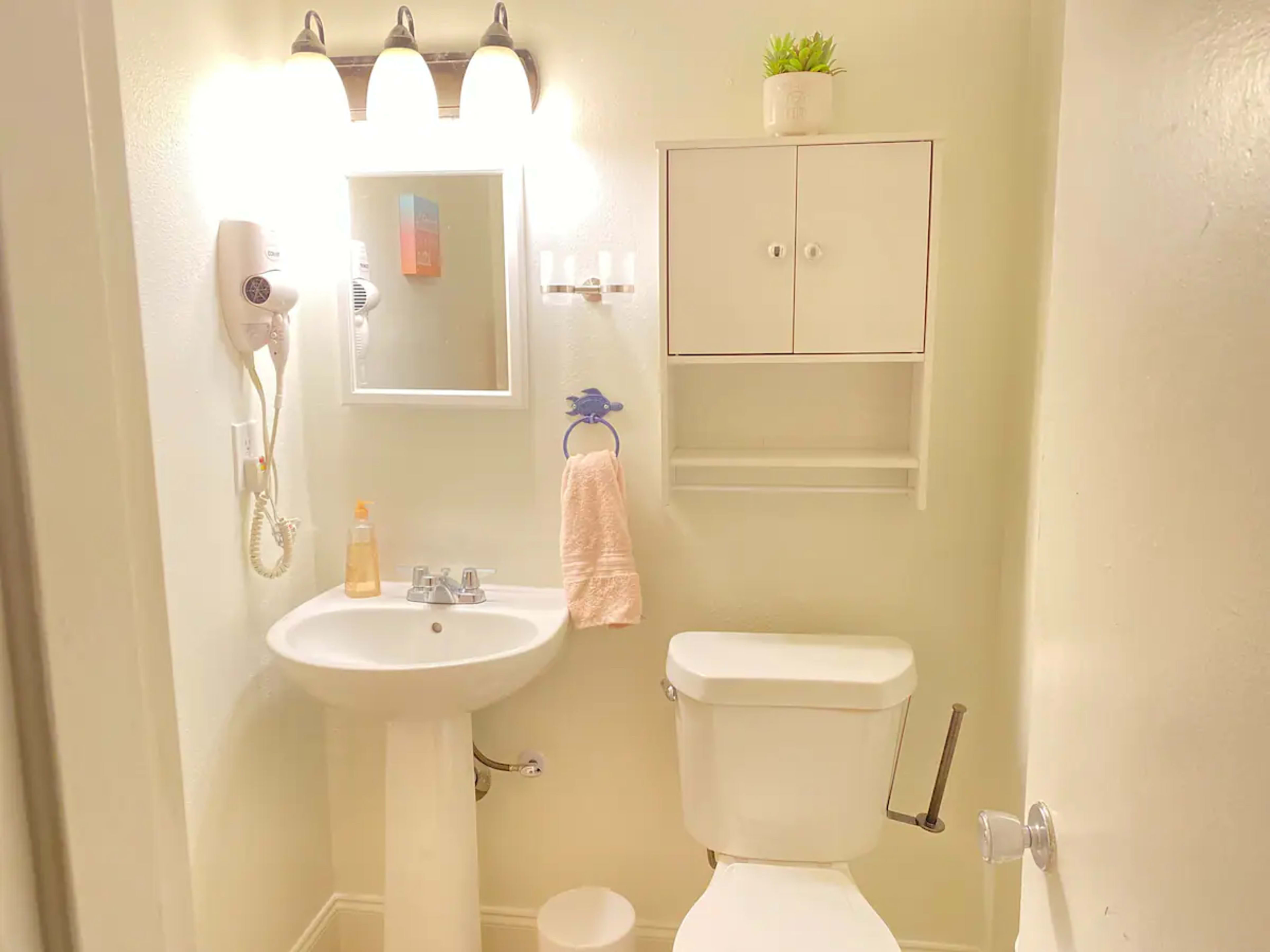 A small bathroom featuring a sink with a mirror above it, a toilet, a wall-mounted shelf, and a hairdryer attached to the wall.